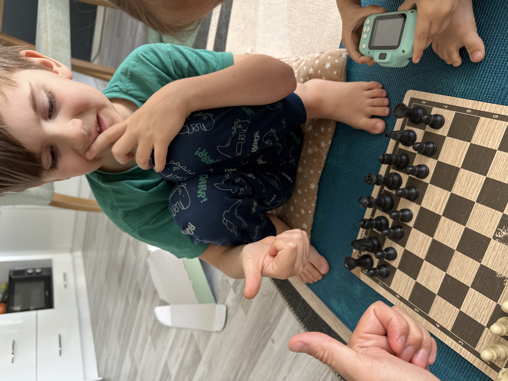 A child wearing a green shirt and dinosaur-patterned pants is sitting on a beige cushion on a blue surface with a chessboard. Black chess pieces are positioned on the board in the upper left corner. A blue smartwatch is in a child's hand in the top right corner. An adult's hand is visible in the bottom right corner, with the thumb extended upward. The room has a gray wood-like floor and white cabinets and appliances are visible in the background. The location is a home interior; there's no indication the city is Fethiye, Turkey.