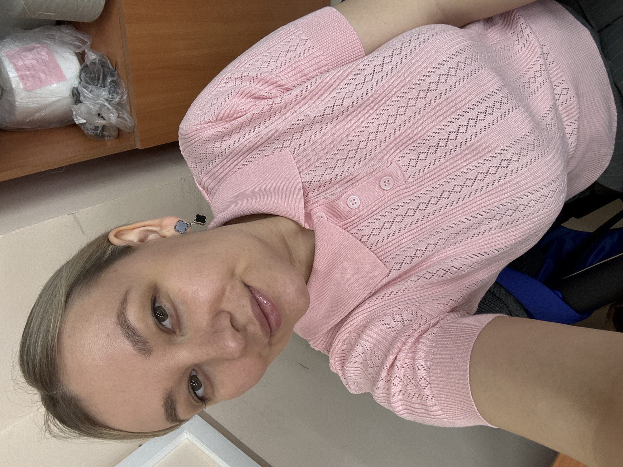 A woman is taking a selfie. She is wearing a light pink knit top with short sleeves and a collar. The top has a textured pattern and three buttons at the collar. She has light skin, brown eyes, and light brown hair pulled back. She is wearing earrings. The background is a plain light wall. To the left is a wooden shelf with a roll of white paper and a plastic bag with dark items inside. The location of this image is not provided in the source data. There is no indication that the location of the image is in Chaikovsky, Russia.