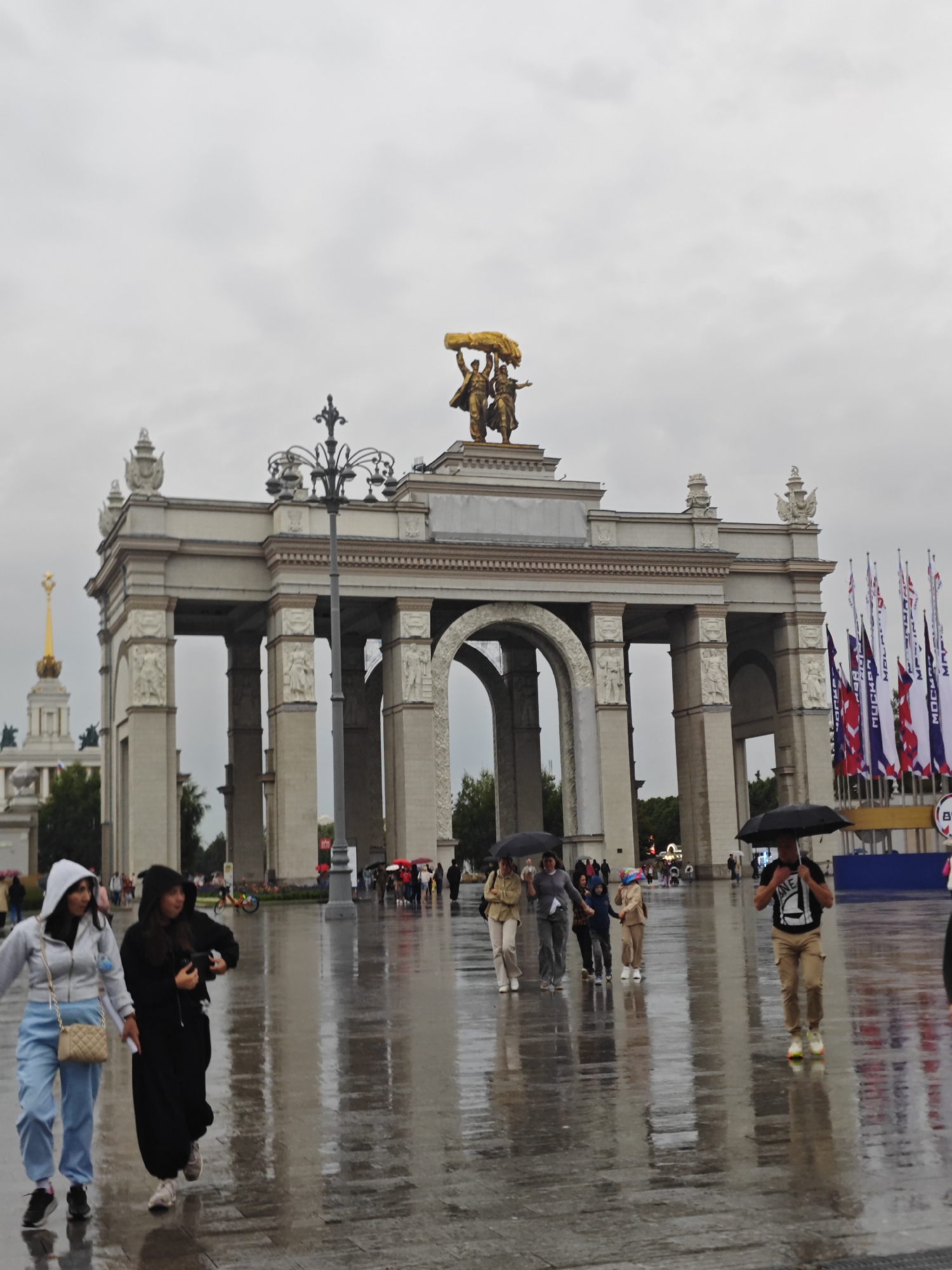The image shows a cloudy day at VDNKh (Exhibition of Achievements of National Economy) in Moscow, Russia. The main subject is the large archway, a prominent landmark at the exhibition center. Several people are walking on the wet pavement, suggesting it has recently rained or is raining. Some individuals are holding umbrellas.
In the foreground, two women are walking from left to right. The woman on the left is wearing a light gray hooded jacket and light blue pants, carrying a beige purse. The woman on the right is wearing a black hooded garment.
Behind them, a group of people are walking towards the right, some with umbrellas. Further back, more people are visible, as well as flags flying on poles.
The archway itself is a grand structure with multiple arches and pillars, decorated with sculptures. On top of the archway, there are golden sculptures of figures. To the left of the main arch, another building with a golden spire is visible. The ground is reflective, indicating it is wet. The overall atmosphere is overcast and damp.
