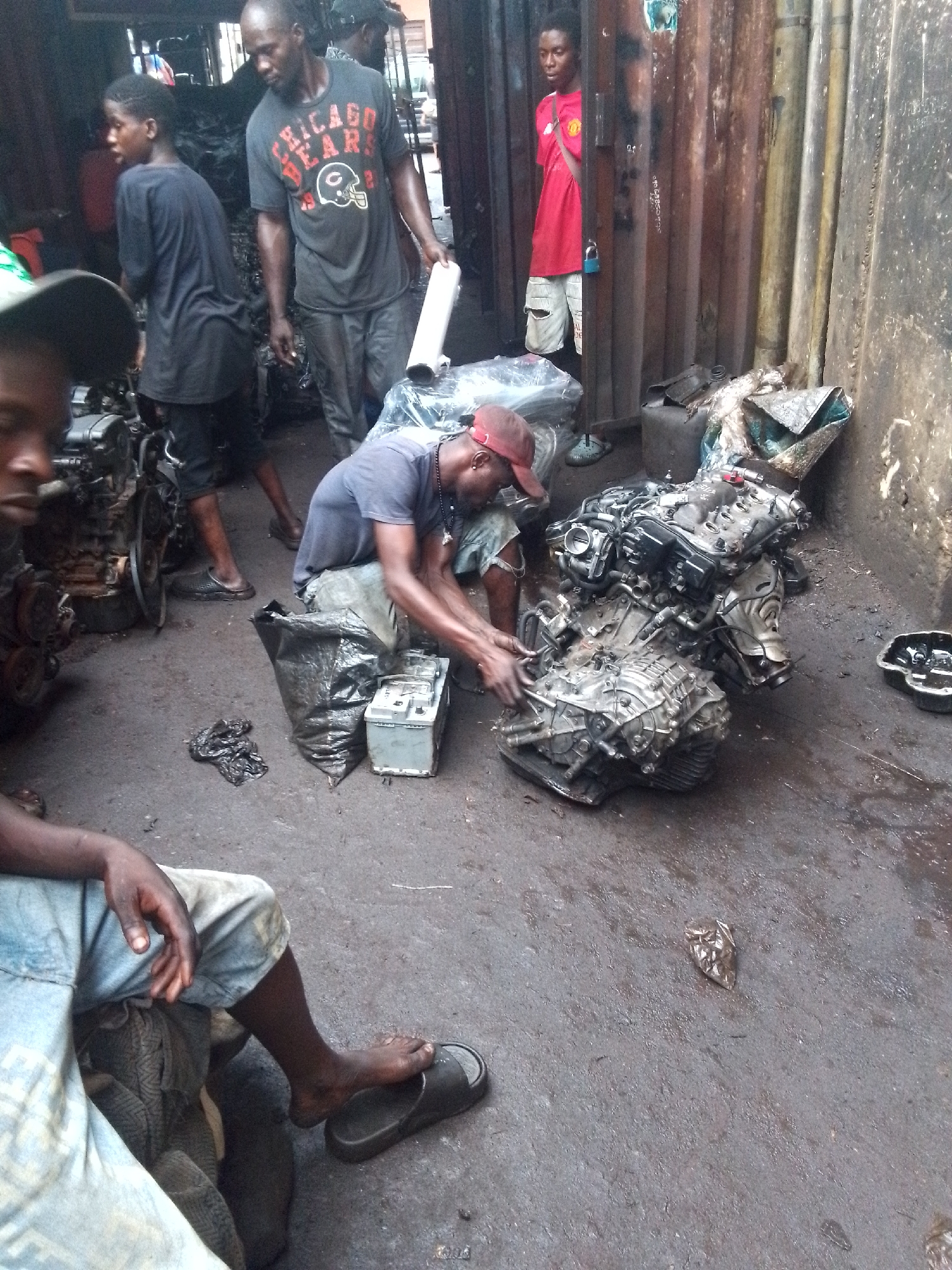 The scene depicts an automotive repair workshop in Onitsha, Nigeria. Several African men are present. One man, wearing a red cap and a dark t-shirt, is working on a car engine and transmission resting on the ground. He is squatting and appears to be manipulating the components. To his left, another man is partially visible, seated and wearing blue denim shorts and a slide sandal. Nearby, another man in a Chicago Bears t-shirt carries a white roll in his left hand, with a youth following behind. In the background, a fourth man stands in the doorway of a corrugated metal structure wearing a red shirt. Car engines and spare parts are scattered around the workshop. The ground appears to be dirt and oily residue.