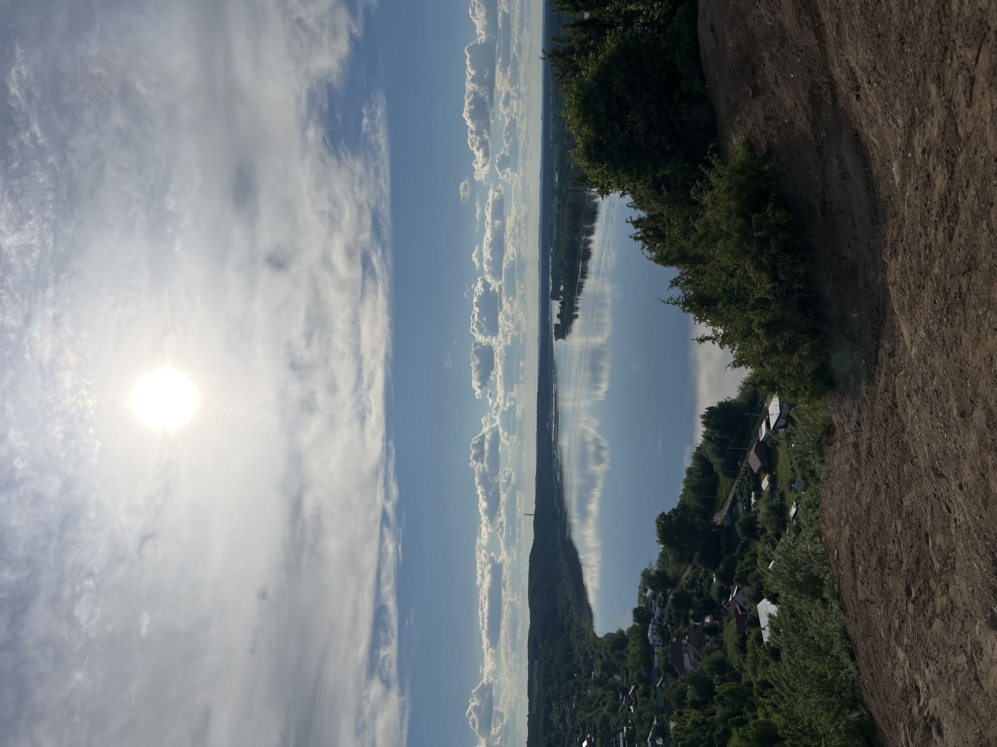 The image presents a panoramic landscape view, likely captured from an elevated position overlooking a wide body of water. The scene is dominated by a clear blue sky, filling the upper portion of the frame, adorned with numerous white clouds ranging from wispy cirrus to more defined cumulus formations. A brilliant, unfiltered sun is visible in the upper left, casting a bright glow that indicates either morning or late afternoon.
Stretching across the middle of the photograph is a broad river or large lake, its calm surface perfectly reflecting the sky and clouds above. The reflections are crisp and elongated, creating an illusion of depth and tranquility. On the far side of the water, a distant landmass or shoreline is visible, appearing low on the horizon.
In the foreground, on the right side, there is a steep, earthy bank covered with sparse vegetation, suggesting a natural incline or a hillside. Below this bank, and along the near side of the water, a verdant landscape unfolds, densely populated with various trees and lush foliage. Nestled among these trees is a settlement featuring several houses and other structures, their roofs and walls peeking through the greenery, indicating a quiet rural or semi-rural community.
There are no people or specific activities discernible in the image. The overall impression is one of peaceful natural beauty and human habitation integrated into the landscape. No visible text is present. The scene depicts a clear day with good visibility. Given the context, the location is Ольховка, Russia.