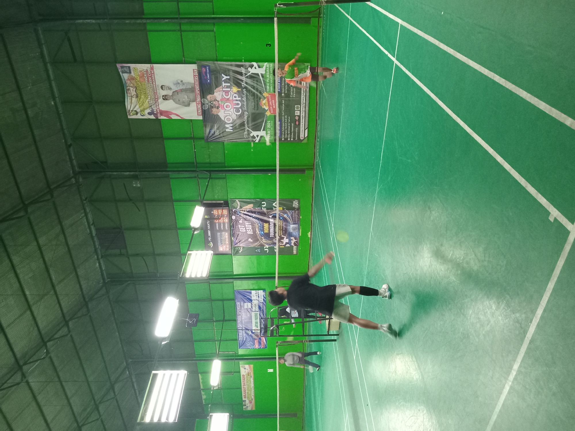 An indoor badminton court in Bojong Gede, Indonesia, is captured from a slightly elevated angle. The court surface is a vibrant green, marked with white lines. Two men are actively playing badminton. One man, wearing a black t-shirt and grey shorts, is in the foreground, mid-swing with a badminton racket, his body blurred by motion. The other man, dressed in an orange and green sports shirt and white shorts, is further back on the court, approaching the net. Two women are also visible on the court, one in the background on the left, appearing to be in a ready stance, and another further behind the net. The court is illuminated by long, fluorescent light fixtures suspended from the green ceiling. Several banners are displayed along the green walls, one advertising a "MOJO CITY CUP" badminton tournament, with dates and sponsorship information visible. Another banner welcomes participants. A scoreboard is mounted on the wall to the right. The overall environment suggests an active sporting event or practice session.