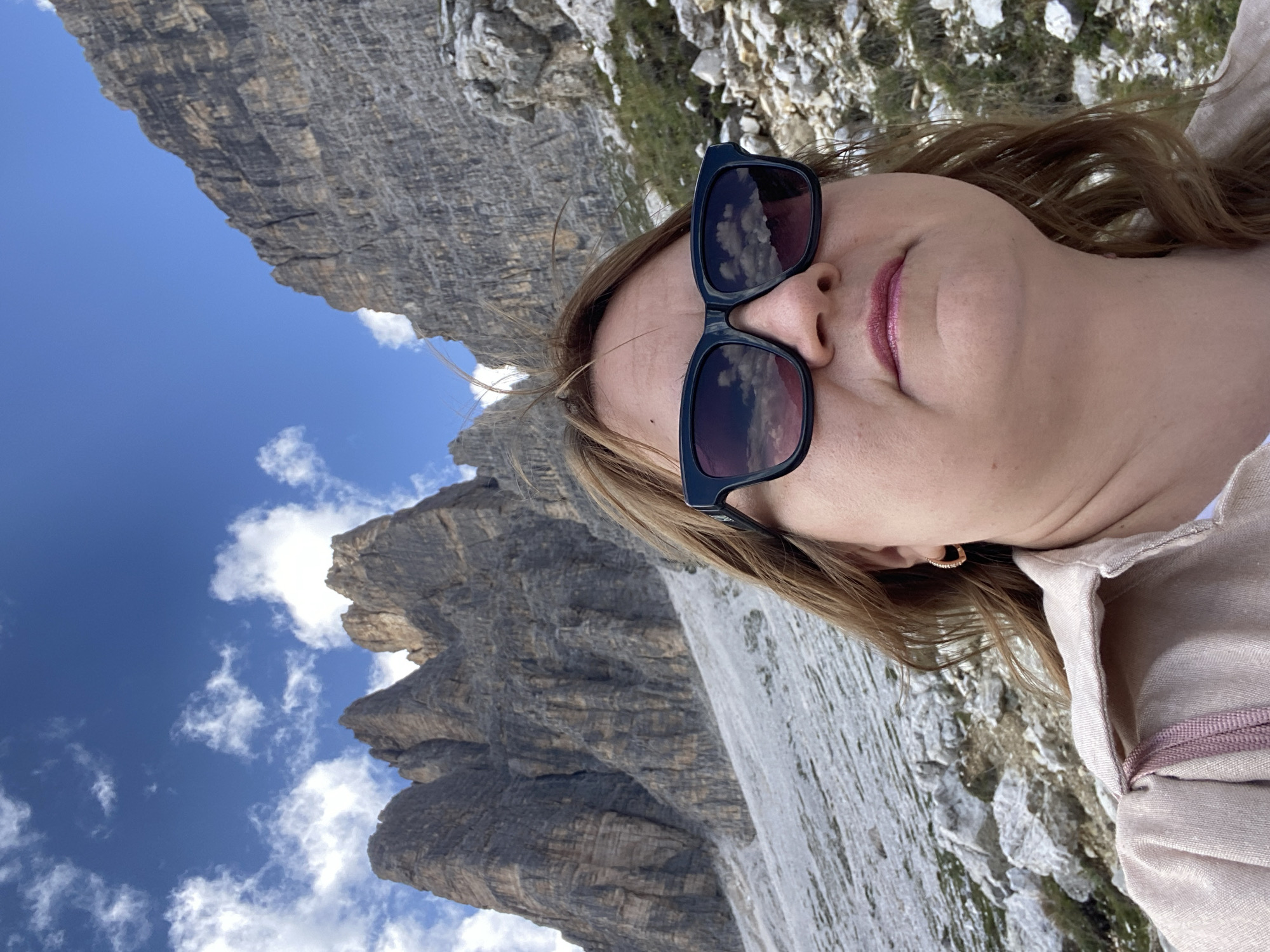 In Auronzo di Cadore, Italy, a woman wearing black sunglasses and a light pink jacket is pictured in a selfie with a mountainous landscape. The woman's head and shoulders are visible, with the focus on her face and the sunglasses, which reflect the cloudy sky and mountains. Behind her, jagged rock formations rise against a bright blue sky dotted with white clouds. The terrain in front of the mountains is rocky and sparse, with patches of dry grass. A pink drawstring from a bag is visible on her shoulder. The image captures a moment during a hike or visit to the mountainous region.