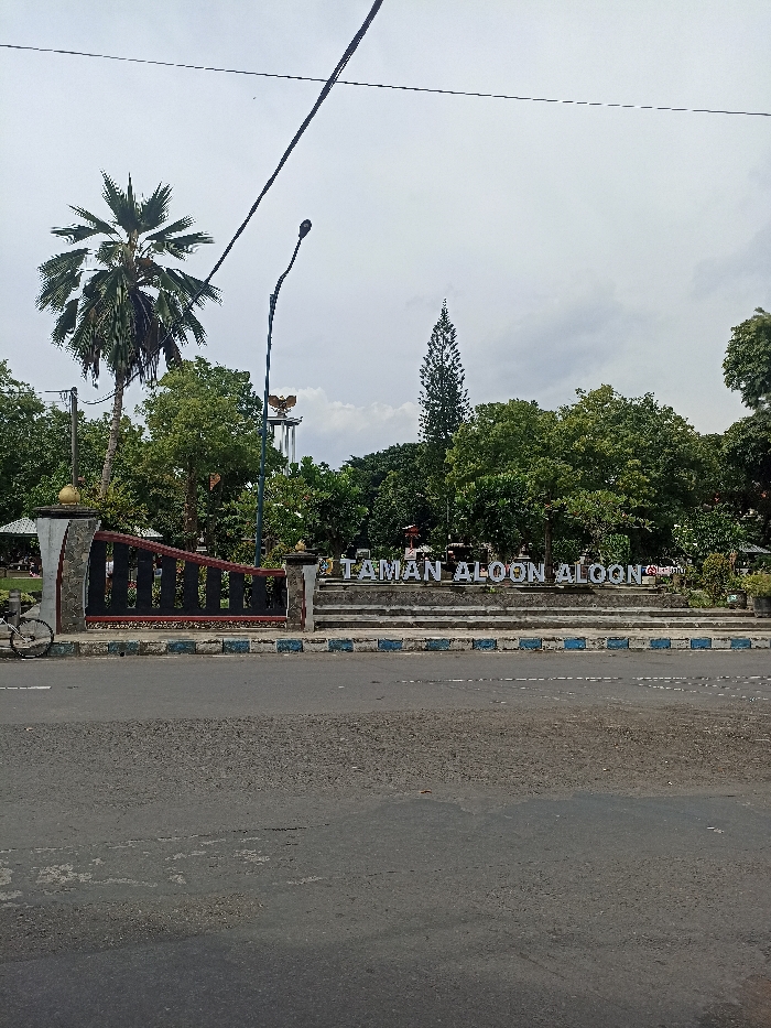A wide-angle outdoor photograph captures the entrance to a public park or square, likely an "Aloon Aloon," situated adjacent to an asphalt road under an overcast sky. In the immediate foreground, a dark gray asphalt road stretches horizontally, delineated by a curb marked with blue and white stripes.
To the left, a portion of a bicycle's front wheel is visible near a decorative stone and dark metal fence, which forms the boundary of the park. Prominently displayed in the midground is a large, white, three-dimensional sign spelling out "TAMAN ALOON ALOON," set on a multi-level stone platform. Behind this sign and the fence, the park is filled with a variety of green trees and dense foliage. A tall palm tree stands out on the left, while a slender evergreen tree rises centrally above the canopy of other deciduous trees.
In the distant background, atop a tall, multi-tiered pedestal, a golden statue, resembling the Garuda Pancasila (the national emblem of Indonesia), can be seen peeking above the treetops. A black street lamp post with a curved arm and a diagonal power line traverse the upper portion of the frame. The sky is predominantly white and cloudy, indicating an overcast daytime. No individuals are present, and no particular events are occurring, portraying a serene view of a public recreational area in what is identifiable as an Indonesian setting, specifically Tulungagung.