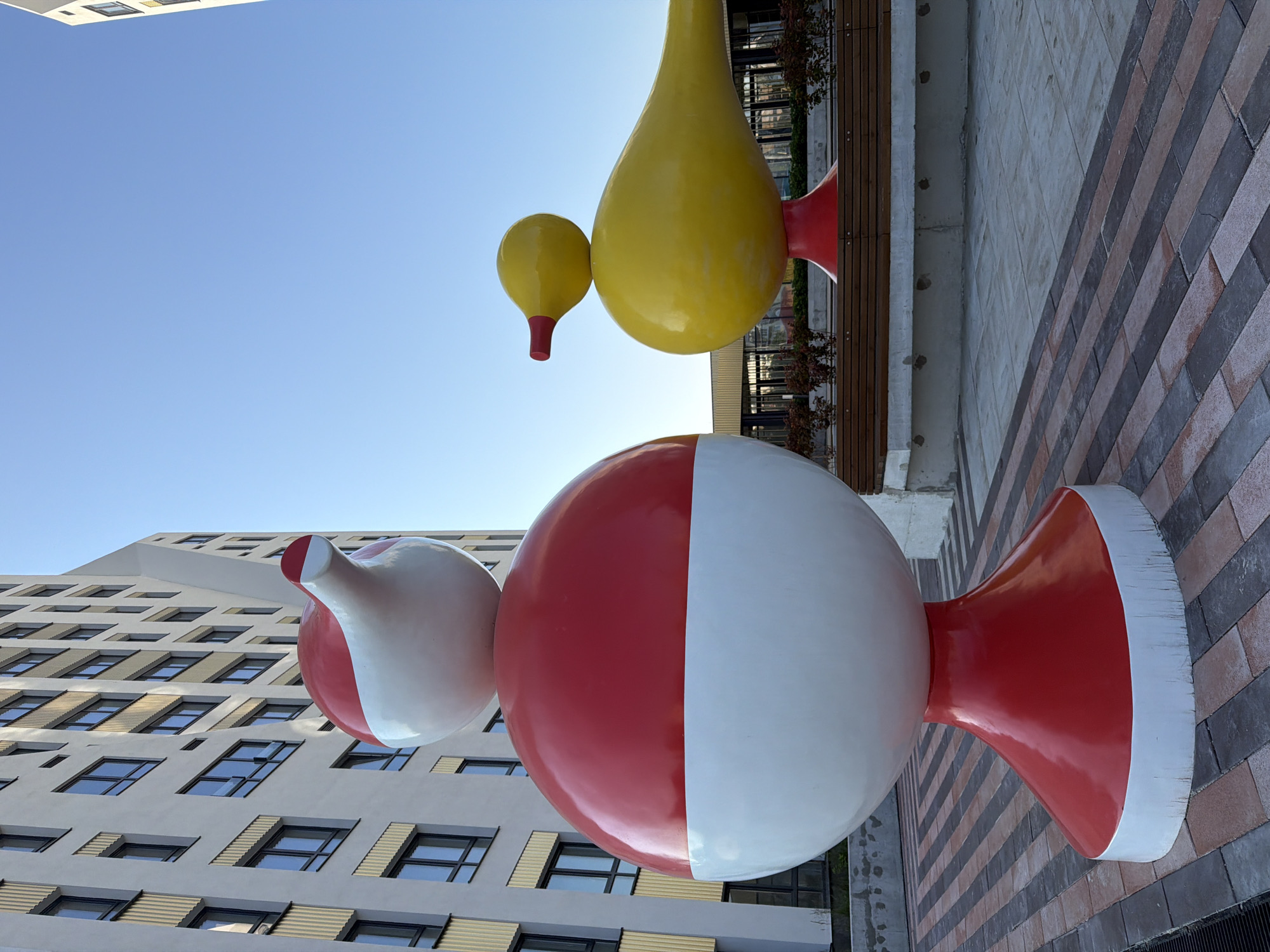 The image shows a modern urban outdoor space in Yekaterinburg, Russia. In the foreground, there is a large, red and white abstract sculpture, resembling a stylized bobber or buoy, composed of stacked spheres and a flared base. To the right, a bright yellow abstract sculpture of a duck, also stylized with a red beak and red base, is prominently displayed. Behind the sculptures, a modern building with a light-colored facade and numerous windows is visible, suggesting a residential or commercial complex. The ground is paved with a pattern of rectangular bricks in shades of grey, brown, and red. A wooden bench with a concrete base sits in the middle ground, with a row of small, dark green bushes planted behind it. The sky is clear and blue, indicating daytime. The scene appears to be clean and well-maintained. There are no people visible in the image.