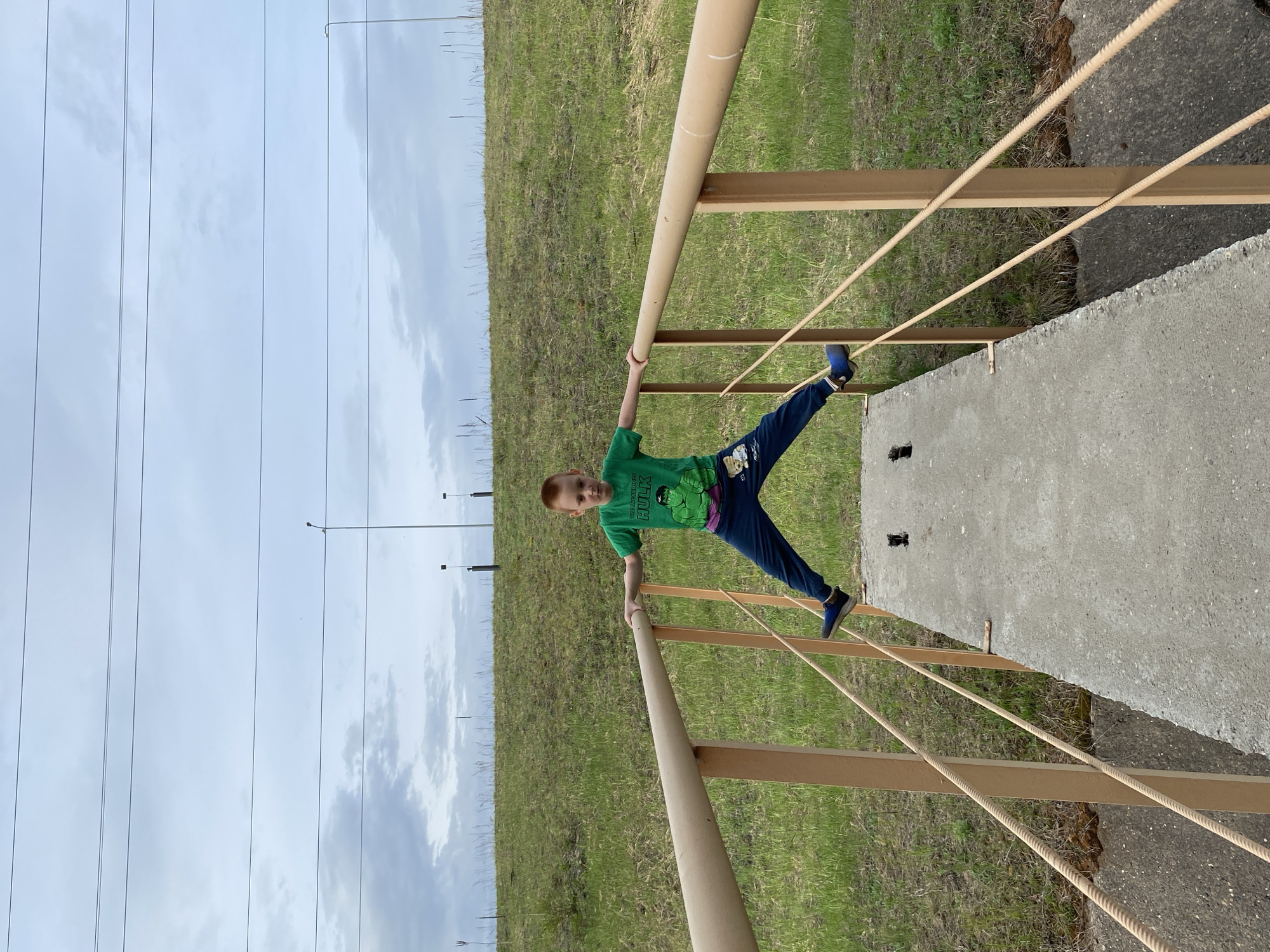 A young boy in a green Hulk t-shirt and blue pants is hanging from a metal railing on a bridge. The railing is painted beige and has rope strung between the top and bottom bars. The bridge appears to be made of concrete. Behind the bridge is a grassy hill. In the background are power lines and utility poles. The sky is overcast. Based on the latitude and longitude, the approximate location is Noviy, Russia.
