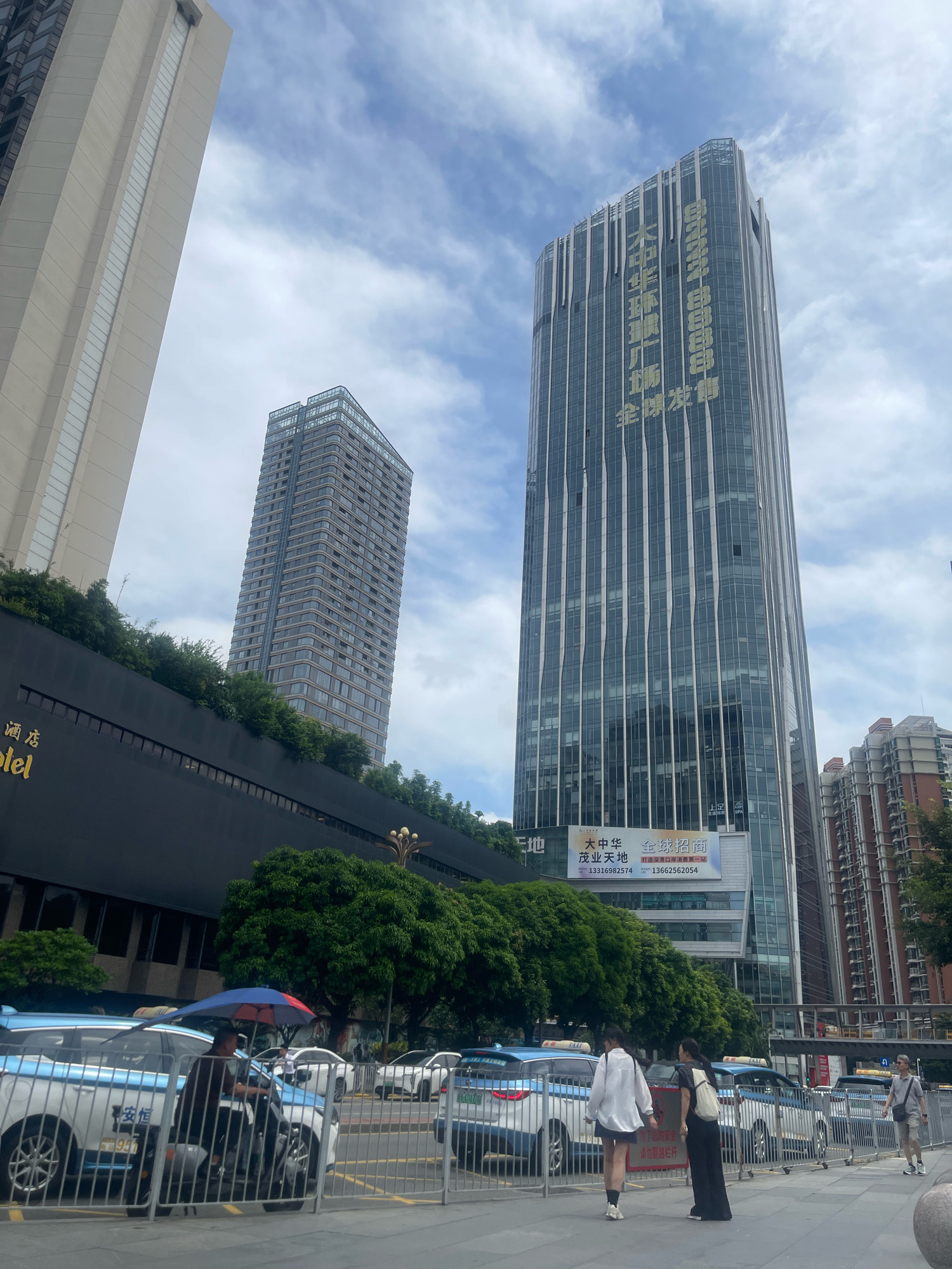 The image depicts a street scene in the Luohu District of China, likely during the day given the illumination and sky conditions. Dominating the background are several modern skyscrapers, characterized by glass facades and vertical architectural elements. The most prominent building on the right features golden vertical text. To the left, a lighter colored, angular skyscraper stands tall. Between these, a mid-rise building with numerous windows is visible. In the foreground, a street is lined with a metal fence and several blue and white taxis, some of which have umbrellas open over them, suggesting a hot or sunny day. Two women, one in a white top and dark skirt and the other in a long black dress, are walking away from the camera along the sidewalk. A man in a light-colored shirt and shorts is walking towards the right side of the frame. Trees and greenery are present along a raised embankment behind the taxis, separating the street from the buildings. A large billboard with Chinese text is visible on the lower part of the taller skyscraper.
