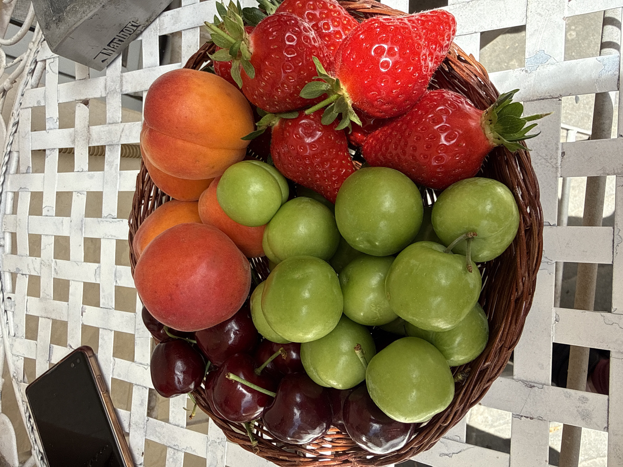A basket of fruit rests on a white, woven table. The fruit includes strawberries, apricots, green plums, and cherries. A smartphone lies on the table near the basket. A metal object with the word "Marlboro" on it is visible in the upper left corner. The location is Sofia, Bulgaria.