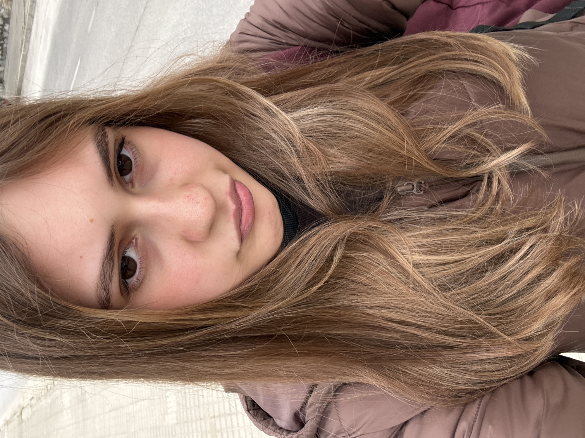 The image is a close-up portrait of a young woman, taken outdoors in what appears to be Panagyurishte, Bulgaria. The photo is oriented vertically, with the woman's head tilted to the right, giving the impression she is lying down and looking upwards, or the photo was taken in landscape and rotated.
The main subject is a young woman with long, wavy, light brown hair that has lighter blonde highlights. Her hair is parted and flows around her shoulders. She has fair skin, visible freckles across her nose and cheeks, and dark brown eyes, which are looking directly at the viewer. Her expression is neutral to slightly smiling. She has natural-looking eyebrows and eyelashes.
She is wearing a dark brown or taupe puffer jacket, which is partially visible, indicating cooler weather conditions. A dark-colored garment is visible underneath the jacket, possibly a turtleneck. The jacket features a zipper on the front.
The background is indistinct, consisting of light-colored, possibly tiled or textured surfaces, suggesting an outdoor setting like a building facade or a wall. There are no specific landmarks or text visible in the image to provide more location context beyond the provided Panagyurishte, Bulgaria. The lighting suggests it is daytime.