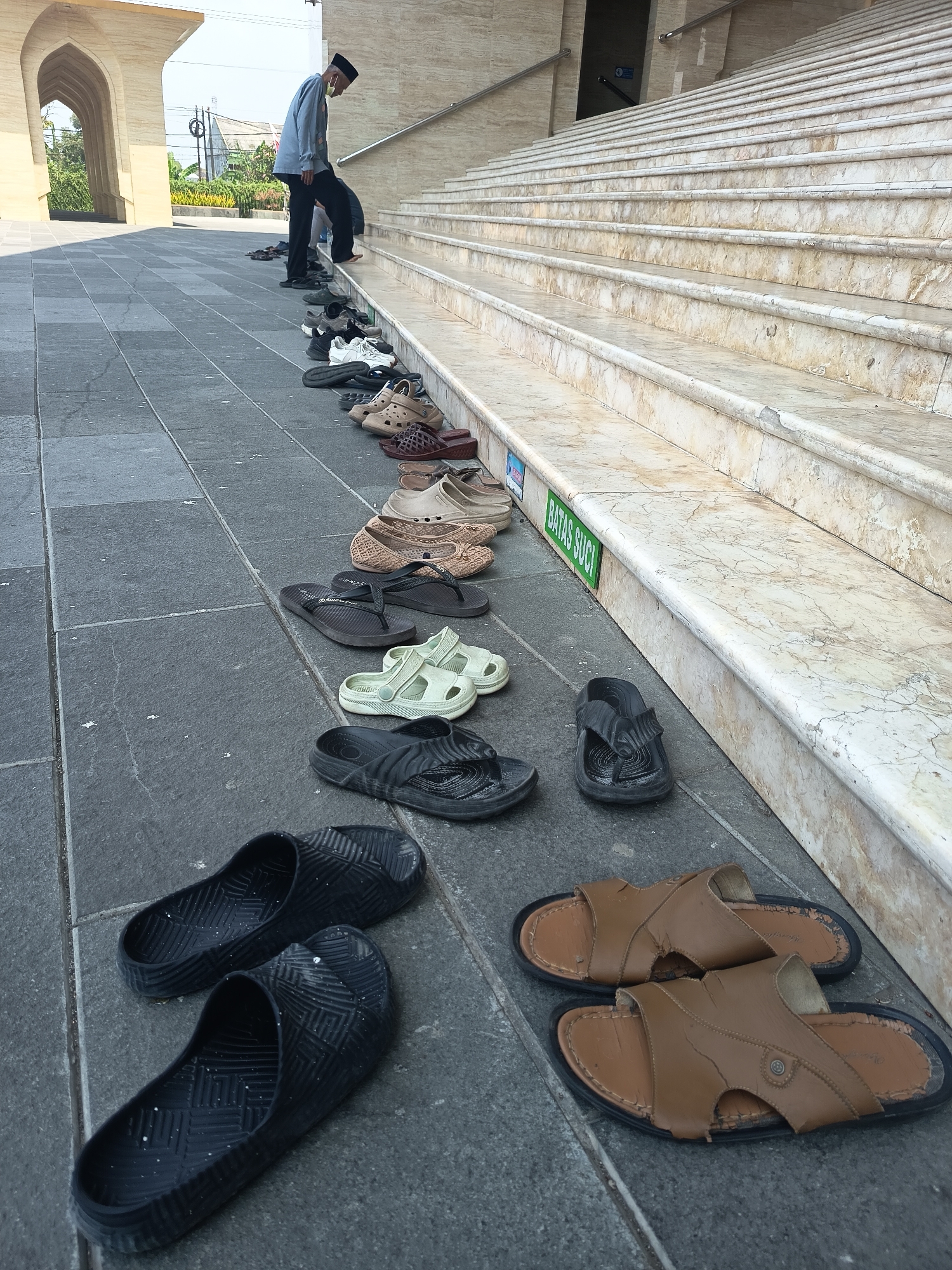 In Dapur, Indonesia, outside a building with tan marble stairs, a person wearing a light blue shirt, black pants, and a white cap is walking up the stairs, leaving behind a line of shoes. The shoes are placed neatly on a dark gray tiled walkway. Among the shoes are several pairs of sandals, including brown leather slides, black flip-flops, and light green slip-on sandals. The area appears to be an outdoor entrance to a religious building, indicated by a green sign on the stairs that reads "BATAS SUCI" (Sacred Boundary). The lighting suggests it is daytime with bright sunlight.