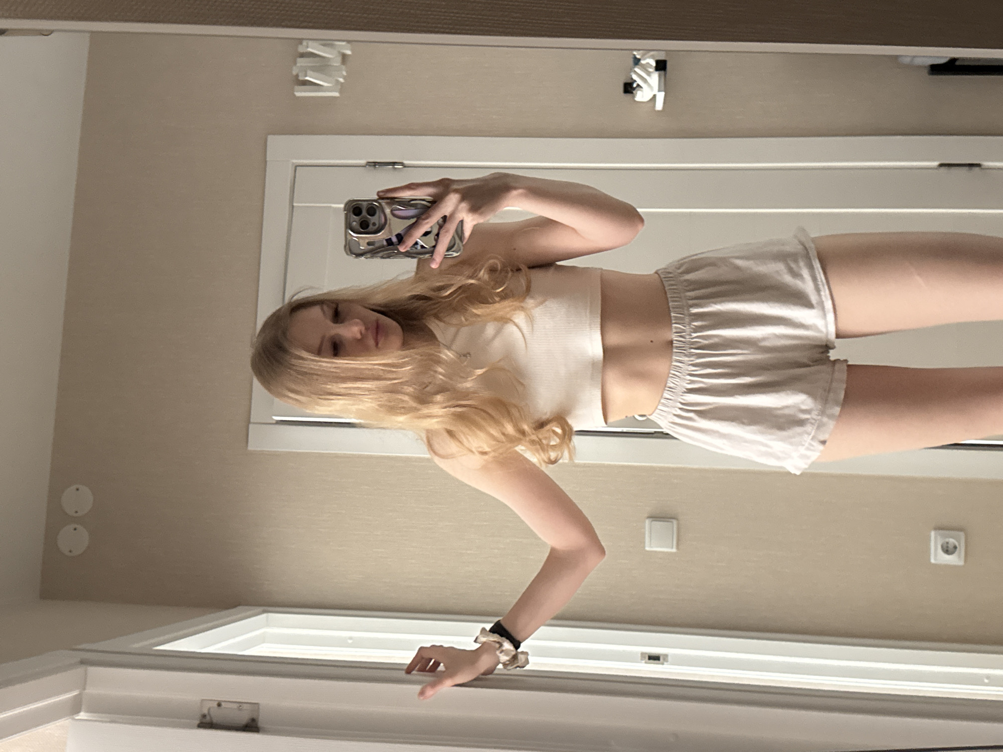 A young woman with long blonde hair is taking a selfie in a mirror. She is wearing a light-colored crop top and shorts. She is holding a smartphone in her right hand, and her left hand is resting on the top of the window frame. The room appears to be a hotel room with beige walls, a white door, and various electrical outlets and fixtures. It is not possible to verify the city as Perm, Russia.