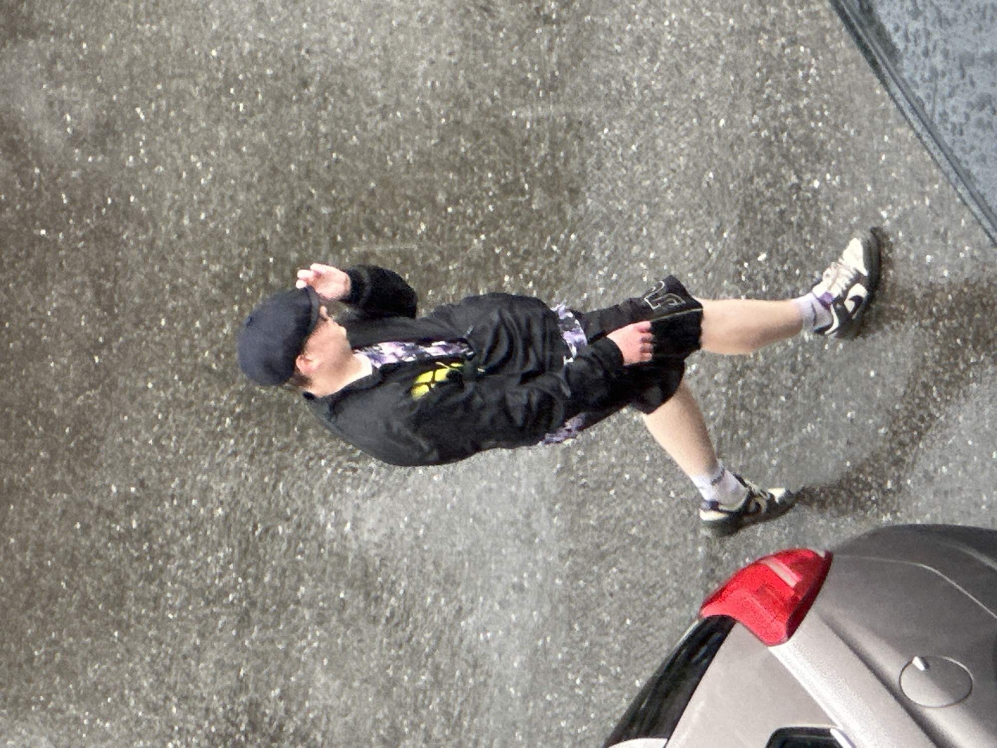 A person is walking on a wet, grey asphalt surface, possibly during rainfall, as evidenced by the speckled appearance of the ground. The individual is wearing a dark flat cap, a black jacket with a colorful, patterned undershirt visible at the neck, black shorts, white socks with purple accents, and dark sneakers with white detailing. The person is holding their left hand up to their cap. To the left of the person is the rear section of a light-colored car, featuring a prominent red taillight. The scene appears to be an outdoor urban or suburban environment. The image composition is an overhead shot. The location is specified as Podolsk, Russia.