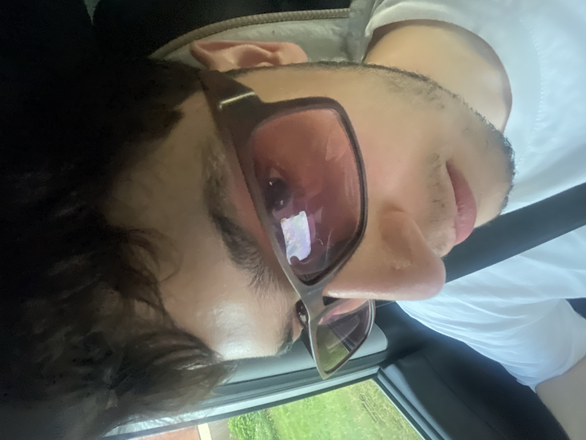 A close-up, low-angle, over-the-shoulder shot shows a young male wearing sunglasses and a white t-shirt. He appears to be in a vehicle, as indicated by a seatbelt strap visible across his chest and a window on the left side of the frame through which green foliage and a building facade can be seen. The lighting suggests it is daytime. The man's face is partially obscured by dark, wavy hair and the large, brown-framed sunglasses with pink lenses. His right ear is visible. The background outside the window appears to be a residential area. The image was taken in Votkinsk, Russia.
