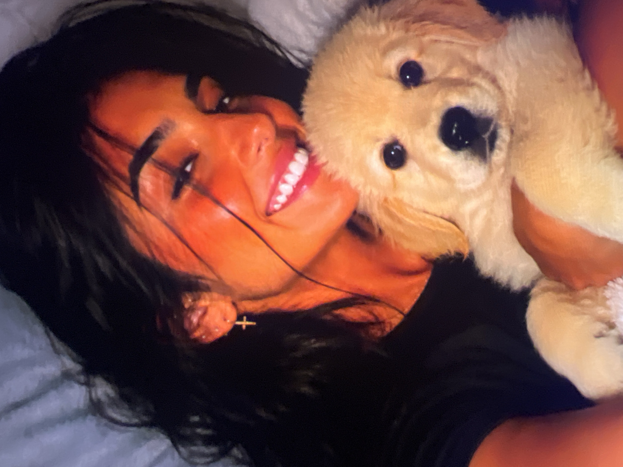 A person, likely a woman with dark hair, is holding a golden retriever puppy in their arms. The woman is smiling and looking towards the camera, with her face illuminated by warm lighting. The puppy's face is visible, with dark eyes and a dark nose. They appear to be indoors, possibly in a bedroom, with soft blankets or bedding visible in the background. The overall scene conveys a sense of warmth and affection. The location is Unknown, Unknown.
