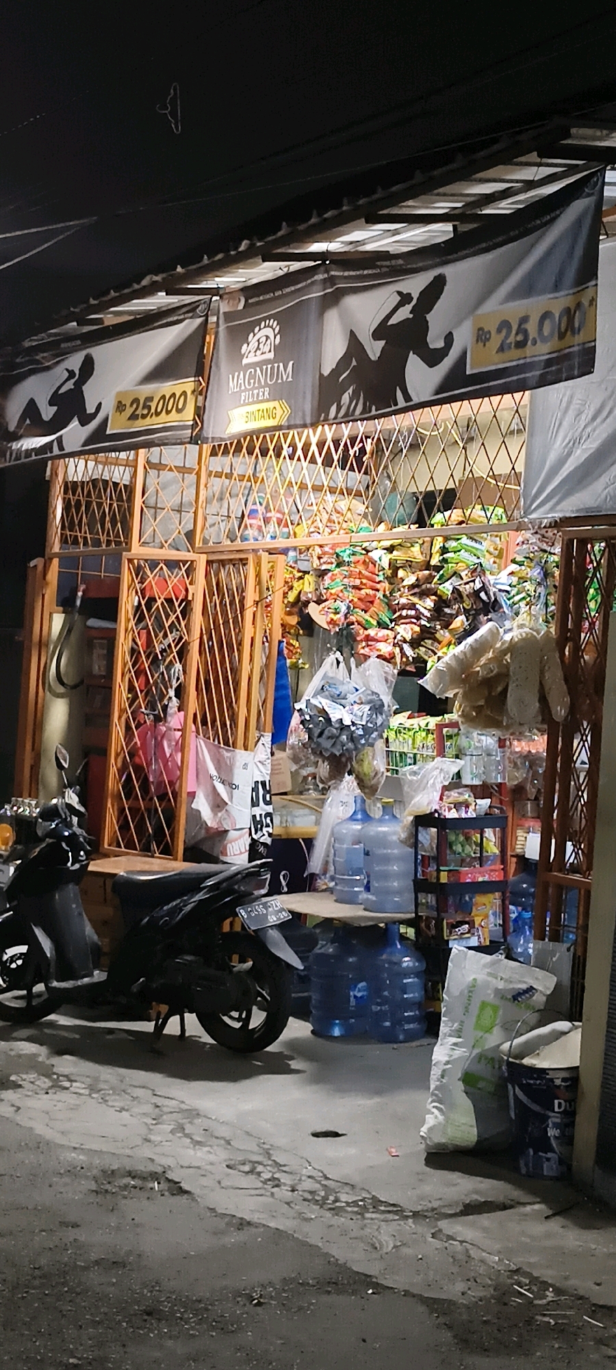 The image shows a storefront at night, likely a convenience store or small warung, in South Tangerang, Indonesia. The storefront is cluttered with merchandise, including stacks of bottled water, bags of snacks, and other items. Two motorcycles are parked in front of the store. A banner above the store advertises "MAGNUM FILTER BINTANG" with a price of "Rp 25.000". The overall impression is one of a busy, informal retail setting.