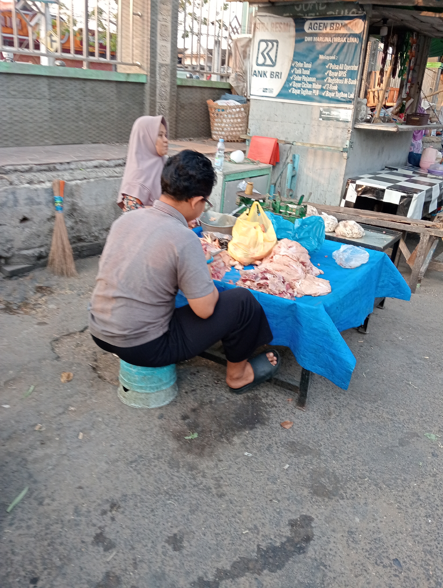 The image depicts an outdoor scene in what appears to be a street or marketplace in Tanubayan, Indonesia. In the foreground, a man wearing a grey t-shirt and black pants sits on a turquoise stool, facing away from the viewer. He is in the process of preparing raw meat, which is piled on a blue tarp spread over a table. Beside him, to his left, a young woman wearing a light purple hijab and a patterned dress is partially visible, also facing away from the camera. A broom with orange and blue accents leans against a concrete barrier behind the woman.
The background features a small roadside kiosk with signage. One prominent sign displays "AGEN BRI" with the logo of Bank Rakyat Indonesia and a woman's name, "DWI MARLINA (MBAK LINA)", followed by a list of services offered, including "Setor Tunai", "Tarik Tunai", "Setor Pinjaman", "Bayar Cicilan Motor", "Pulsa All Operator", "Bayar BPJS", "Registrasi M-Bank", "T-Bank", "Bayar Tagihan PLN", and "Bayar Tagihan Listrik". To the right of the kiosk, a checkered black and white fabric covers a surface.
The environment is paved with asphalt, showing some scattered debris and water stains. The lighting suggests it might be late afternoon or early morning, with soft sunlight casting shadows. The overall impression is a mundane street-side commercial activity.