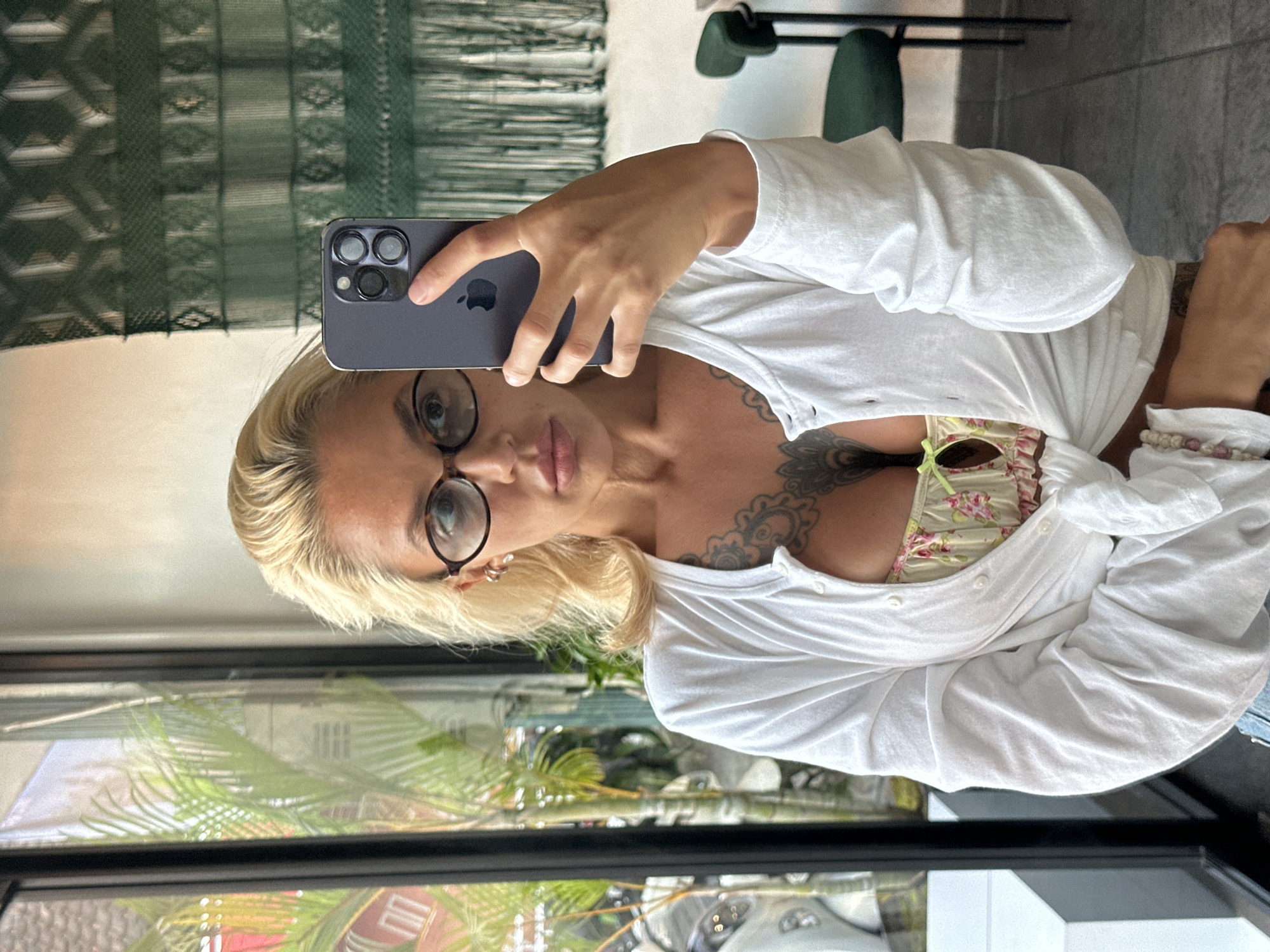 A woman with blonde hair and glasses takes a selfie in a well-lit indoor setting. She is wearing a white open cardigan over a floral-patterned top, with visible tattoos on her chest. The background shows a large window with greenery outside, a tapestry hanging on the wall, and a dark green chair. The woman is holding a smartphone to capture the image. The scene appears to be a cafe or restaurant.