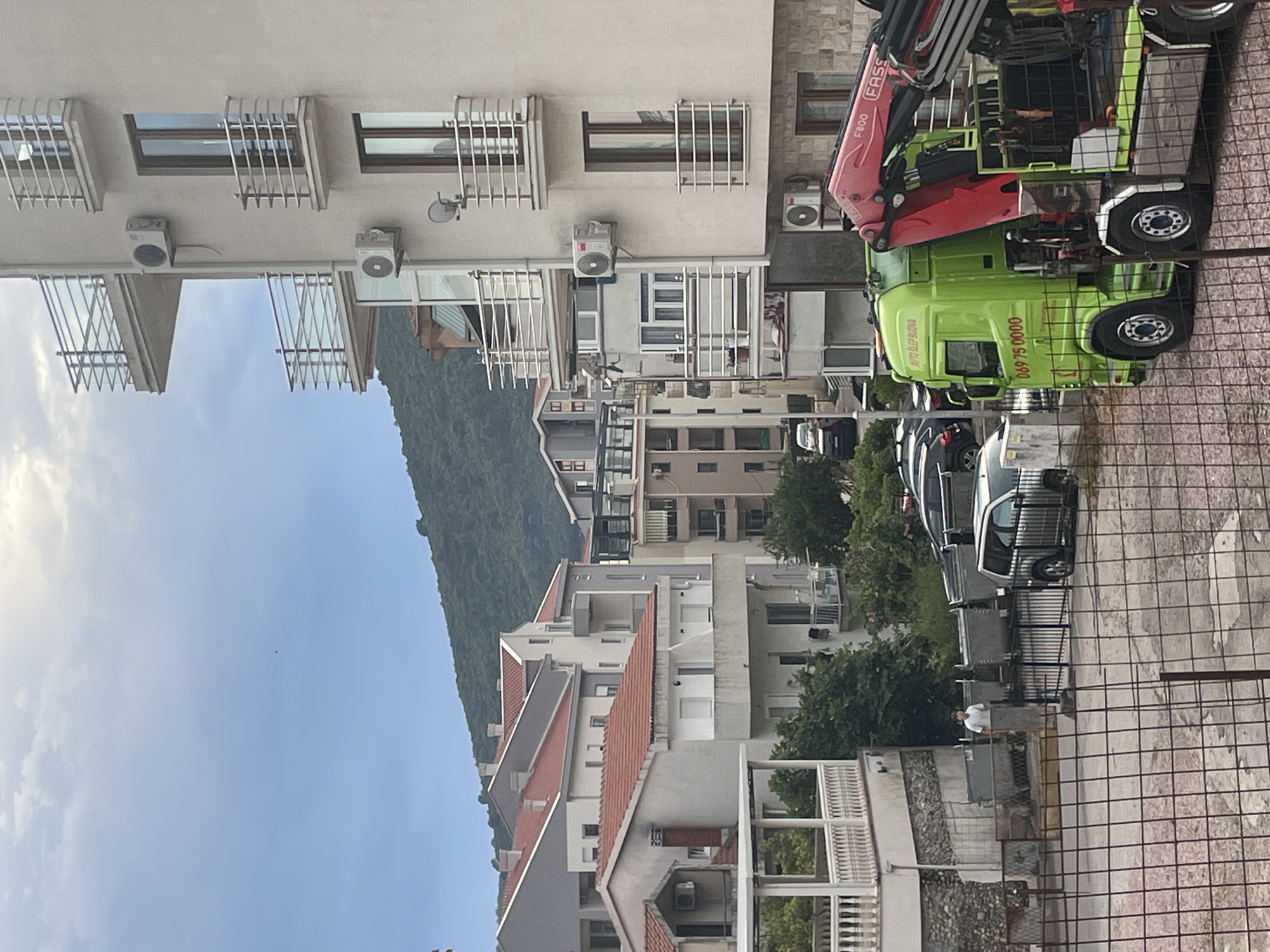 The image captures a street scene in Budva, Montenegro, with a focus on a bright green truck equipped with a crane, parked on the right side of the frame. The truck has "AUTO SLEP BUDVA" and a phone number "069 75 0000" visible on its side. In the background, multiple multi-story buildings with red-tiled roofs and balconies with metal railings are visible. A lush, green hillside forms the backdrop to the urban landscape. In the foreground, a man in a white t-shirt stands near a fence, and several large dumpsters are also present. Cars are parked along the street and on a slight incline in the distance. The sky is overcast with some clouds.