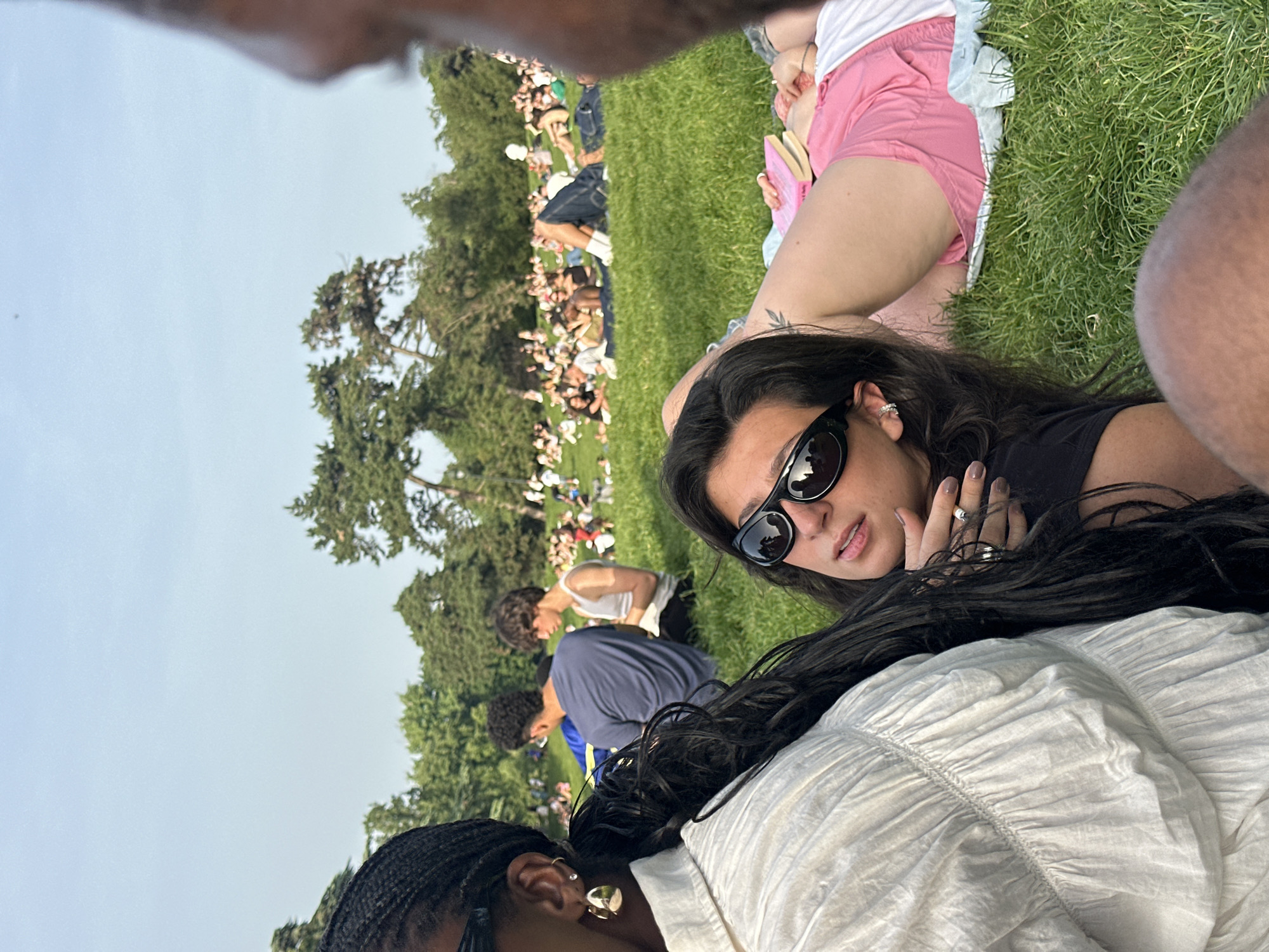 The image captures a scene of people relaxing in a park, likely in Paris, France, based on the casual atmosphere and style. Several people are sprawled out on the grass, including a woman wearing sunglasses and a black top lying down. Near her is another woman with braided hair and gold earrings. In the background, a crowd of people is dispersed across the lawn amidst trees. The photo seems candid, possibly taken from the perspective of someone also lying on the grass.