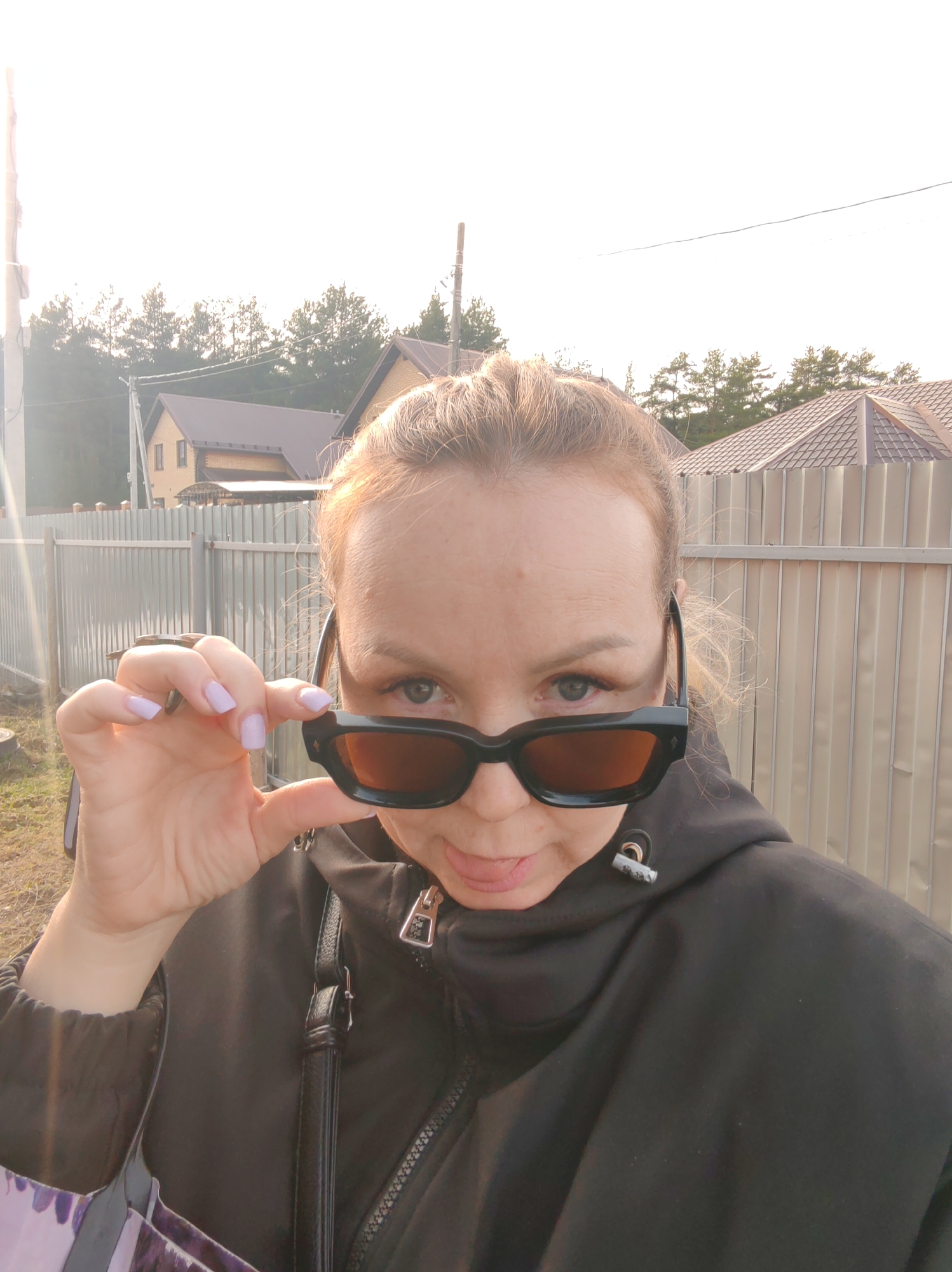 A woman in a black jacket and sunglasses stands outdoors. She is holding the sunglasses up to her face. The background includes a metal fence, houses with brown roofs, and trees. The location is likely in Chaikovsky, Russia, based on prior information.