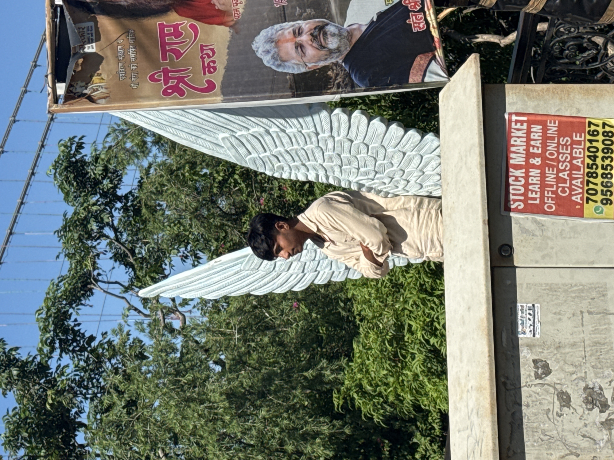 A young man stands with his arms crossed in front of large, light blue angel wings, likely a prop or statue. He is wearing a light-colored, long-sleeved shirt. Behind him, there is a banner that appears to be advertising a religious event, possibly a "Shri Ram Katha," with text in Hindi. A man with a beard and glasses is pictured on the banner. The scene is outdoors, with lush green trees and foliage in the background, and a clear blue sky above. A power infrastructure box or similar structure is visible in the foreground, with a sign advertising "STOCK MARKET LEARN & EARN OFFLINE / ONLINE CLASSES AVAILABLE" with phone numbers. The overall impression is a street scene with a mix of cultural and commercial elements. The presence of the "Shri Ram Katha" banner and the general environment suggest this location is likely in Rishikesh, India, a city known for its spiritual and religious significance.