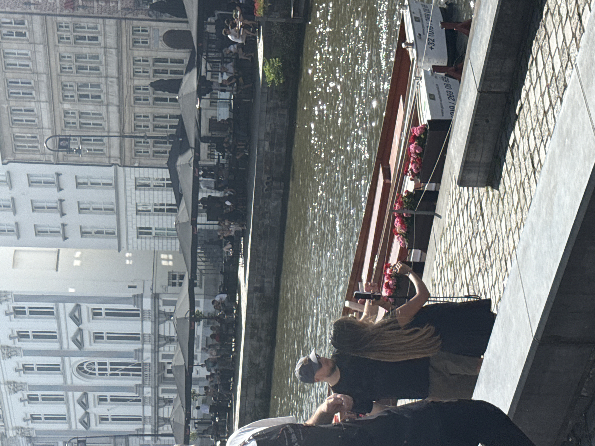 The image shows a sunny day scene along a canal, likely in Ghent, Belgium. In the foreground, two people are visible, one wearing a baseball cap and facing away from the camera. The other person has long blonde hair and is holding a phone, possibly taking a picture or video. They are standing on a cobblestone walkway next to the canal.
The canal is filled with shimmering sunlight reflecting off the water. A boat is moored along the side of the canal, decorated with pink flowers in planters. The boat has text on its side, including "+32 (0)9 269 08 69" and "ghent-waterfront".
Across the canal, several buildings with white facades and numerous windows are visible. In the background, people can be seen sitting at outdoor tables, suggesting a cafe or restaurant setting. The architecture of the buildings, with their repeating window patterns and ornate details, is characteristic of European cities. The overall impression is a vibrant, urban waterside environment on a clear, bright day.