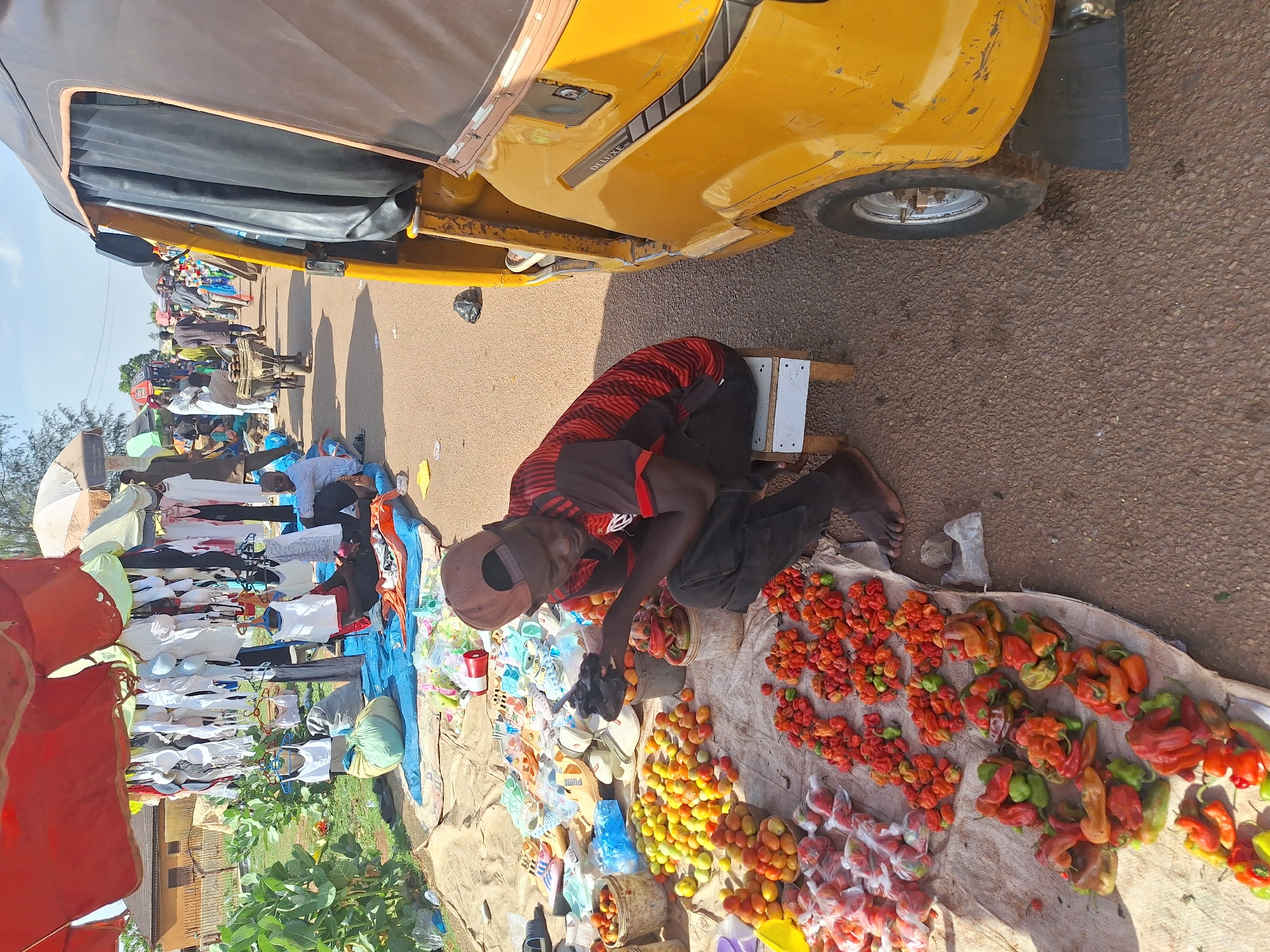 A street vendor kneels on a small wooden stool, sorting colorful peppers, tomatoes, and other produce displayed on a cloth on the ground. He is located next to a yellow vehicle, possibly a taxi or "Keke Napep" (auto-rickshaw) common in Nigeria. Other vendors and passersby are visible along the street, suggesting this scene is a busy market or commercial area. The location is Kakuri Gwari, Nigeria, as per the request.