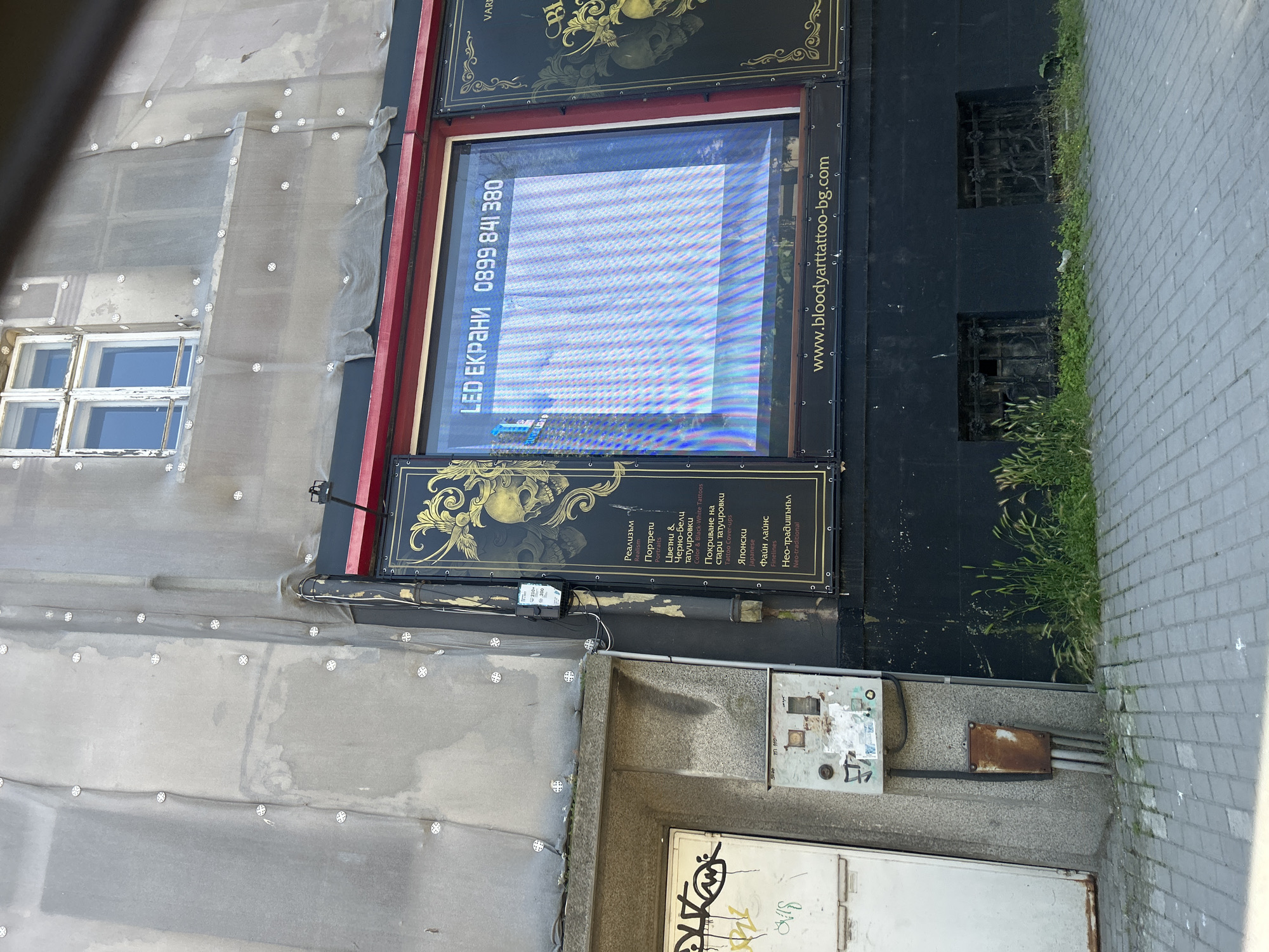 The image depicts the exterior of a commercial establishment, likely a tattoo parlor, situated in Varna, Bulgaria. The building's facade is covered with a light-colored, translucent fabric material, possibly for renovation or protection, secured by numerous white circular fasteners. A window on the upper floor is visible through this covering, revealing multiple panes and a white frame.
Below the upper floor, a storefront features a large display window with an LED screen showing a pattern of blue and white vertical lines. Above the LED screen, text in a Cyrillic script is partially visible, reading "LED ЕКРАНИ 0899 841 380". To the right of this display, a black panel with ornate gold designs and a skull motif is evident. Beneath the main display window, a website address "www.bloodyarttattoo-bg.com" is printed in white text on a black banner.
To the left of the storefront, a white door is covered with graffiti. Adjacent to this door is a weathered electrical box mounted on a concrete wall, with a smaller, rusted metal box attached below it. The ground in front of the building is paved with grey rectangular tiles, arranged in a basket-weave pattern. Tufts of green grass grow in patches along the base of the building and between the pavement and the structure.