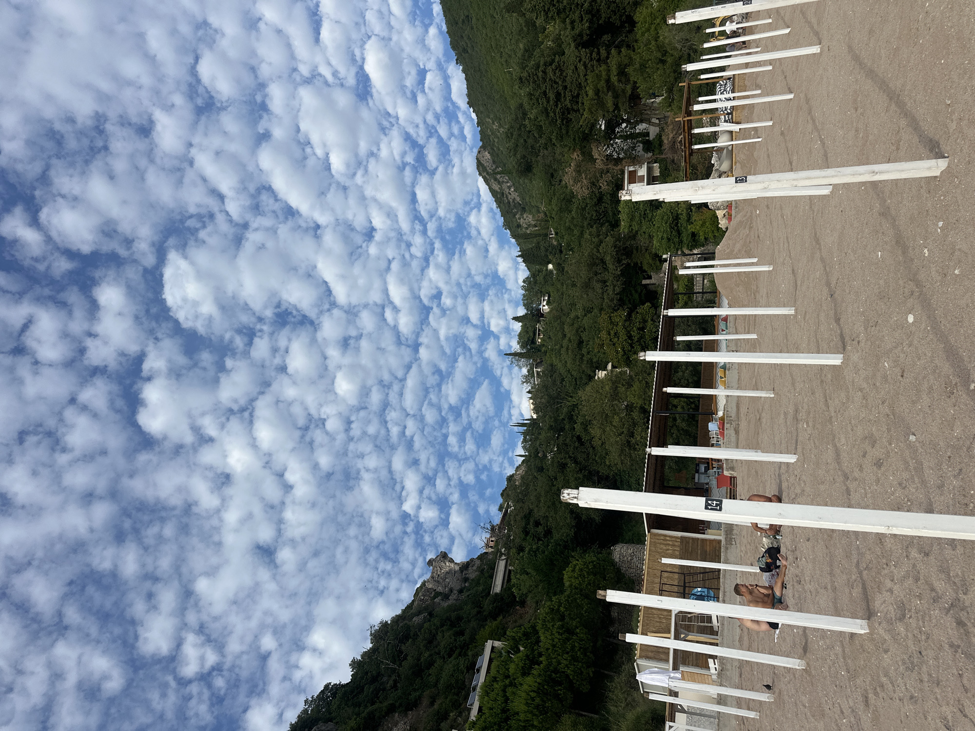 The image presents a wide view of a sandy beach located in Perazića Do, Montenegro, bordered by steep, densely forested mountains on both sides. Above, a bright blue sky is largely covered by a consistent pattern of small, white cumulus clouds.
In the foreground, the beach consists of light brown, coarse sand. Numerous white, rectangular wooden posts are vertically embedded into the sand, extending from the foreground into the mid-ground. Some of these posts are marked with black numbers, notably "14" and "13". Three adult males are seated directly on the sand in the mid-ground, positioned around one of the numbered posts. One male, wearing dark shorts, is oriented facing right. A second male, in light-colored swim trunks, sits opposite a dark backpack. The third male, also in swim trunks, is seated to the right of the group, facing left. A small, dark-feathered bird stands on the sand near the seated individuals.
Behind the men, along the beach line, several temporary or permanent structures are visible. These include various wooden frameworks, a building with a thatched or reed roof, and a white shirt draped over a white wooden frame. Further along the beach to the right, what appears to be construction equipment, possibly a small excavator, is present.
The mountains flanking the beach rise steeply, covered in dense, dark green vegetation comprising numerous trees and shrubs. Rocky outcrops are visible on the left mountain, where scattered residential buildings are nestled into the slopes. One building features solar panels on its roof. Further buildings are discernible within the tree line on both mountain sides.
