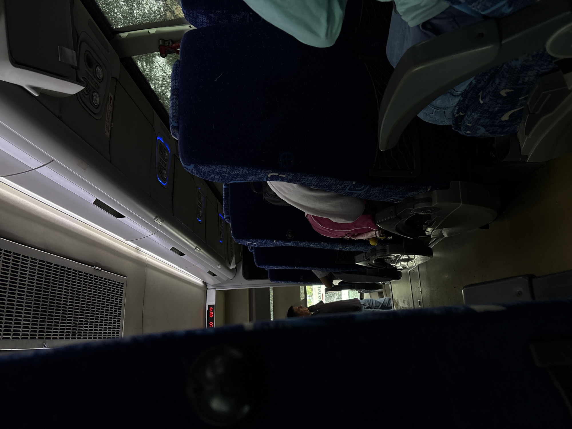 The image shows the interior of a bus, likely in motion or stopped at a station, with multiple rows of blue upholstered seats visible. A portion of a person's arm, clad in a light blue garment, is in the foreground on the right. Behind it, another person's head and shoulder, wearing a light pink garment, are partially visible. In the distance, a man in a dark collared shirt and blue jeans stands near the entrance or exit of the bus. Above the passengers, there are overhead compartments and lighting fixtures. A digital display shows "10 66°F". The windows show a blurred, outdoor environment, suggesting the bus is traveling. There are no specific landmarks or city details visible in the image that would definitively place it in Chilpancingo, Mexico.