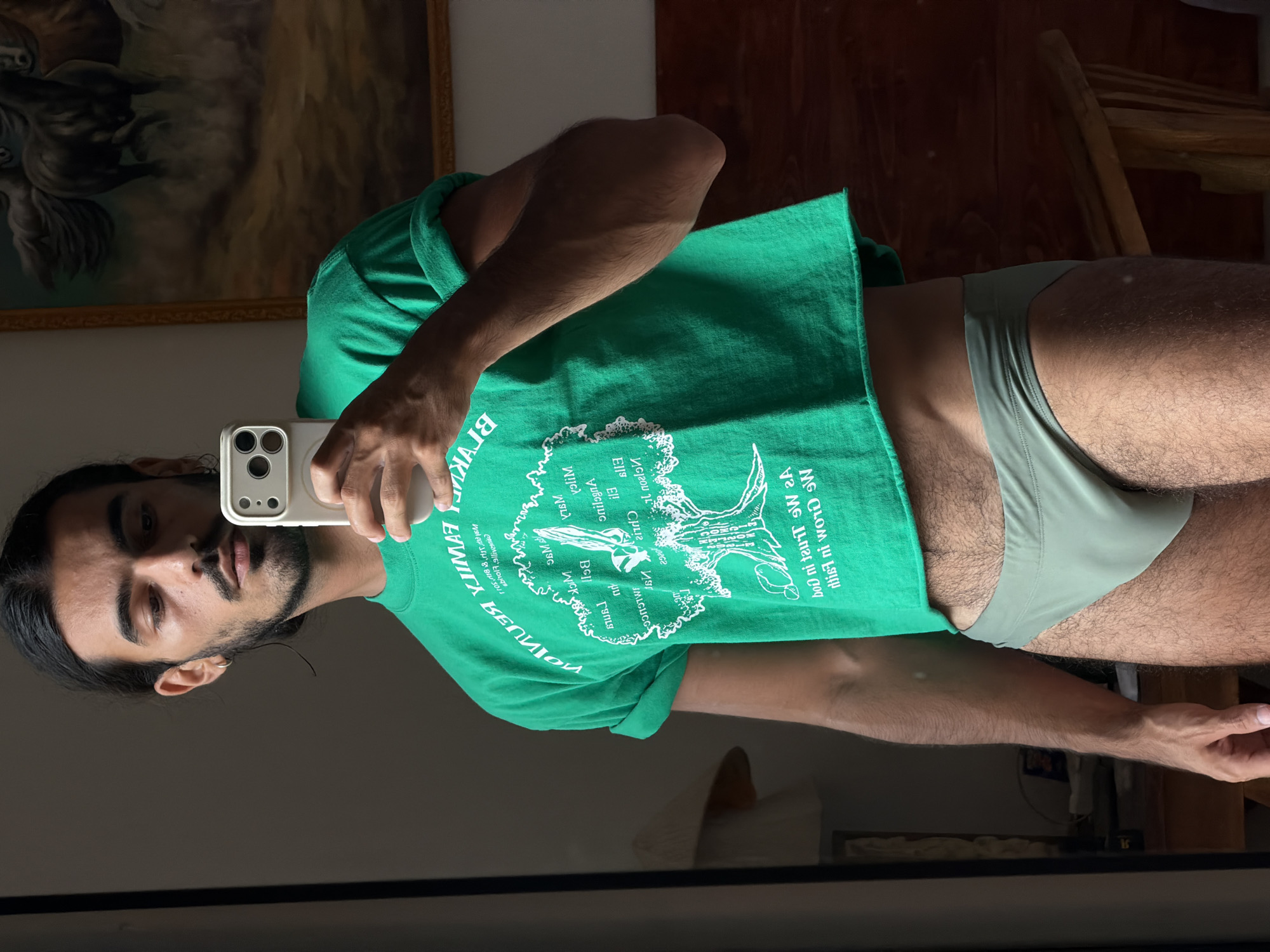 A man stands in front of a mirror, holding a white smartphone in his right hand to take a selfie. He has dark hair pulled back in a ponytail, a dark mustache and beard. He wears a green t-shirt with white text, and light green briefs. In the background is a wall, wooden chair, and framed painting.