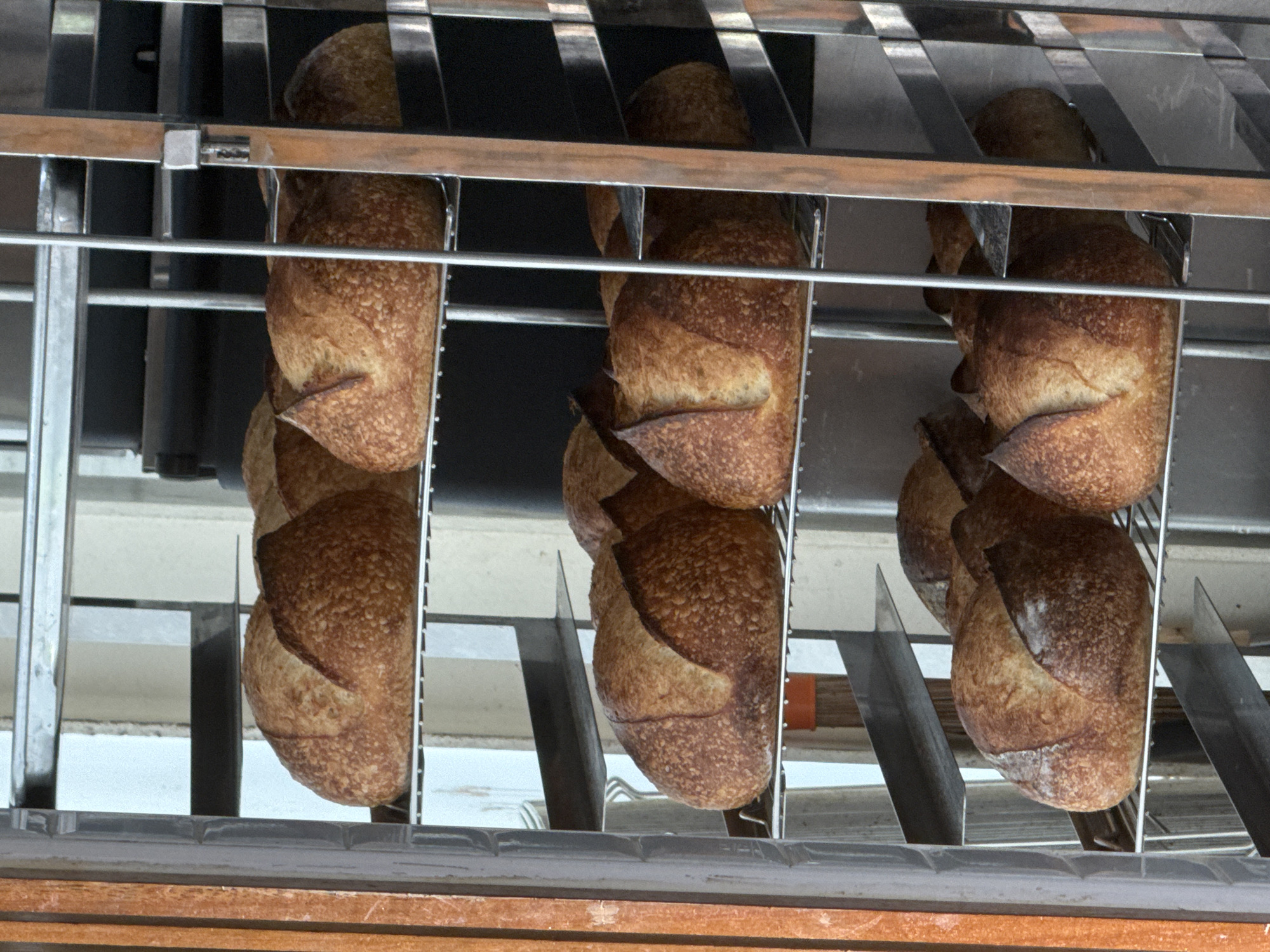 A tall metal rack with multiple shelves is filled with baked loaves of bread. The bread is round with a scored top crust and a golden-brown exterior. The rack is positioned in what appears to be a commercial kitchen or bakery. The background is indistinct, with some shelving and dark surfaces visible. The lighting suggests an indoor environment. This scene is located in Bangkok, Thailand.