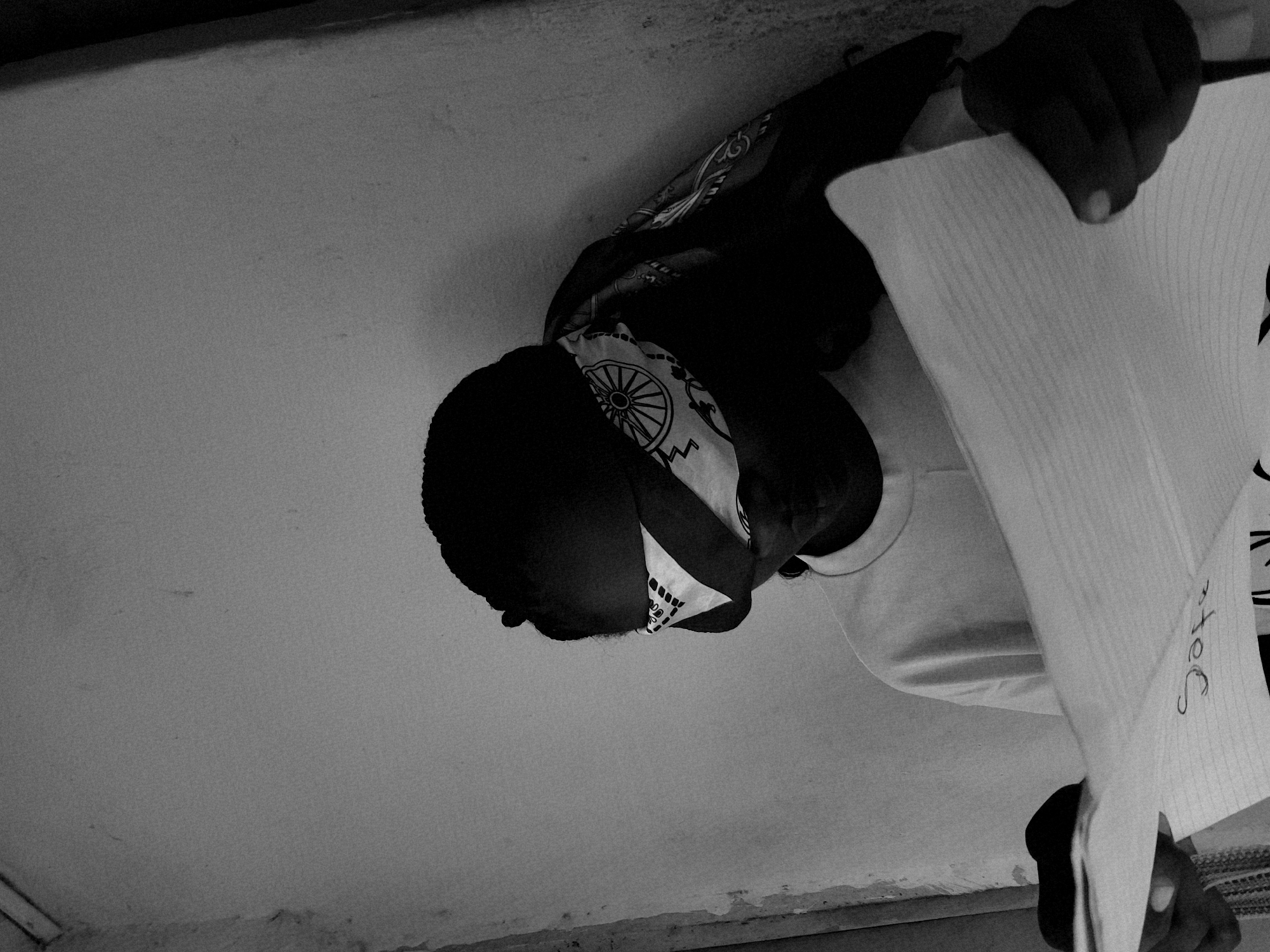 A black and white, low-angle shot depicts an individual holding lined paper with writing on it. The individual appears to be wearing a bandana covering the forehead. The room seems sparsely decorated. The location is possibly Jos, Nigeria.