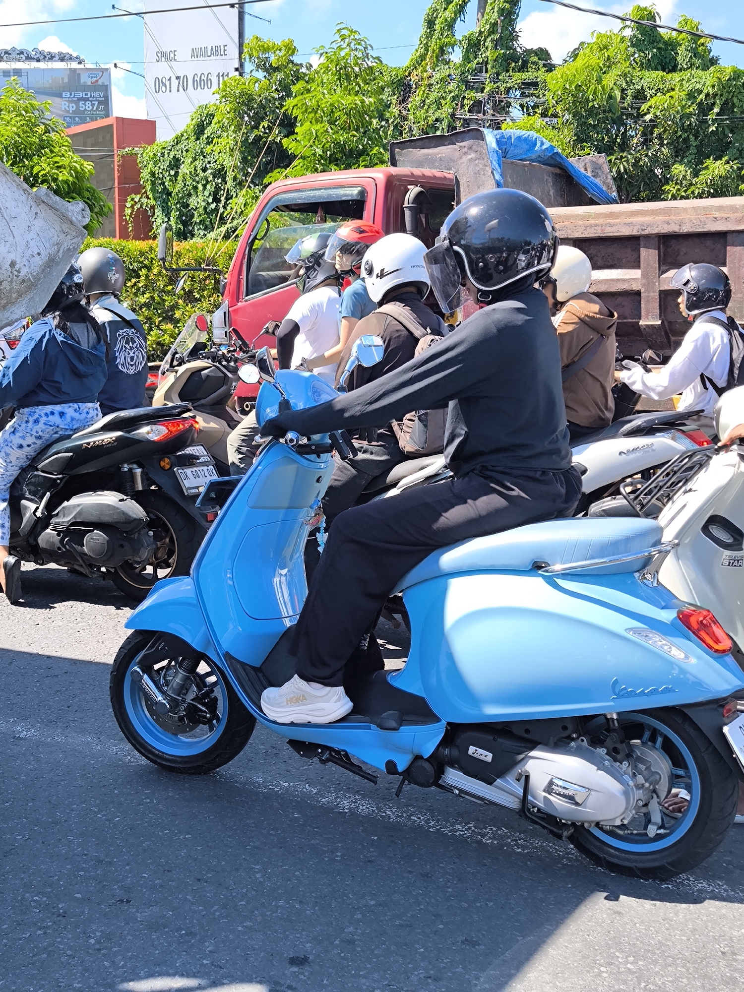 This bustling outdoor scene, set in what appears to be a sunny day in Kuta, Indonesia, captures a street full of stopped traffic. The foreground is dominated by a person seated on a bright blue Vespa scooter, facing right, wearing a black helmet, black long-sleeved shirt, and black pants. Their white sneakers prominently display the "HOKA" brand. The scooter features matching blue wheel rims and a chrome grab handle on the rear seat.
Behind the central figure, a line of other motorcyclists and a large red dump truck are halted. Most individuals are wearing helmets of various colors, including black, white, and red, obscuring their faces. One person on a black Yamaha NMAX scooter has a distinctive lion emblem on the back of their jacket, and the scooter's license plate reads "DK 6012 OK." Another white scooter has "TELEVIS STAR" partially visible on its side, and a white helmet bears the "HONDA" logo. The red dump truck has a rusty bed with a blue tarp covering its contents.
The setting is characterized by lush green trees and overgrown foliage in the background, suggesting a tropical environment. Overhead, wires are visible. Two billboards stand out: one large white billboard proclaims "SPACE AVAILABLE" with the phone number "081 70 666 11," while a smaller billboard mentions "BJ30 HEV" and "Rp 587 JT." The strong sunlight creates clear shadows on the asphalt road, indicating mid-day. The overall activity depicts a common urban scenario of vehicles waiting in traffic.