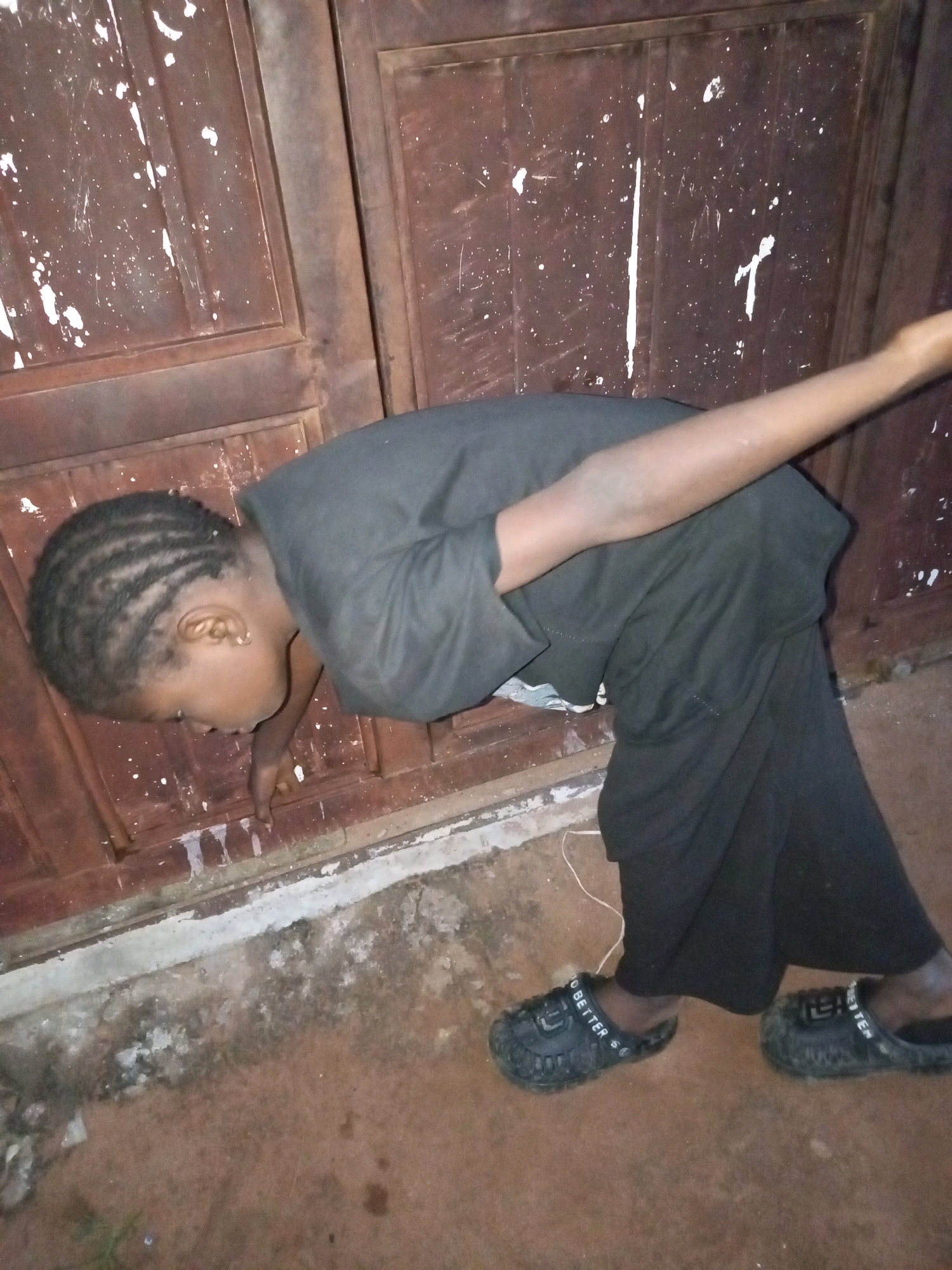 A young person is bending over in front of a dark brown, weathered wooden door, with their right arm extended forward. The door has white paint splatters and vertical marks. The person is wearing a black t-shirt, black pants, and black slip-on shoes with the word "BETTER" visible on the side of one shoe. Their hair is styled in cornrows. The ground in front of the door is a mix of concrete and dirt, with some debris. A thin white wire is visible on the ground. The scene appears to be outdoors, with the lighting suggesting it might be evening or night. There is no clear indication of weather conditions. The location is identified as Tempure, Nigeria.