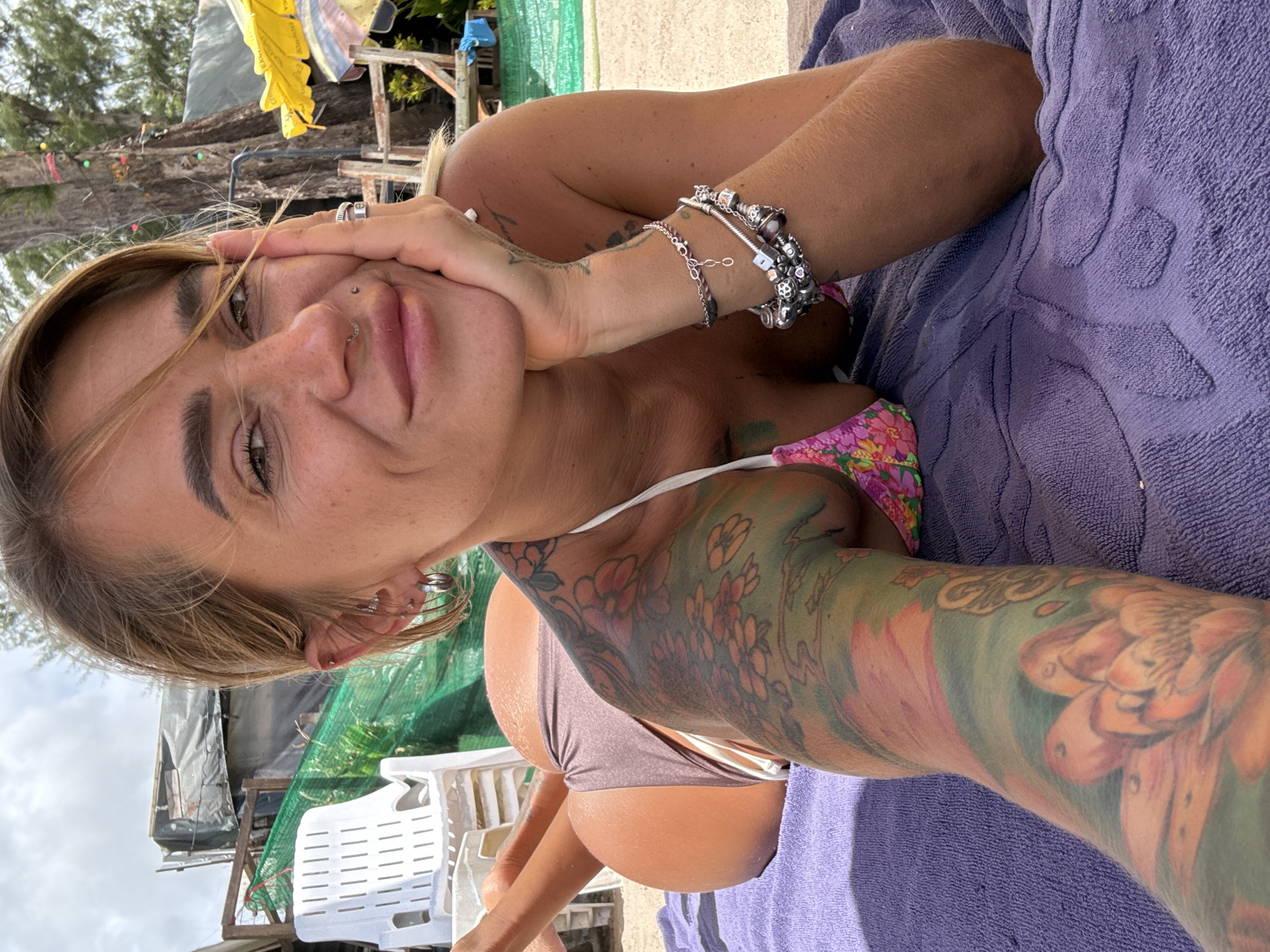 A woman with blonde hair and numerous tattoos, including a floral design on her arm, lies on a purple towel. She is wearing a bikini top with a floral pattern and a solid color bottom. Her hand is resting on her face, and she appears to be looking directly at the camera. She has a nose ring and several rings on her fingers. In the background, there is a green net structure, some trees, and what appears to be part of a building or shelter. The scene suggests an outdoor setting, possibly a beach or resort area, given the towel and the tropical foliage. The lighting indicates it is daytime.
