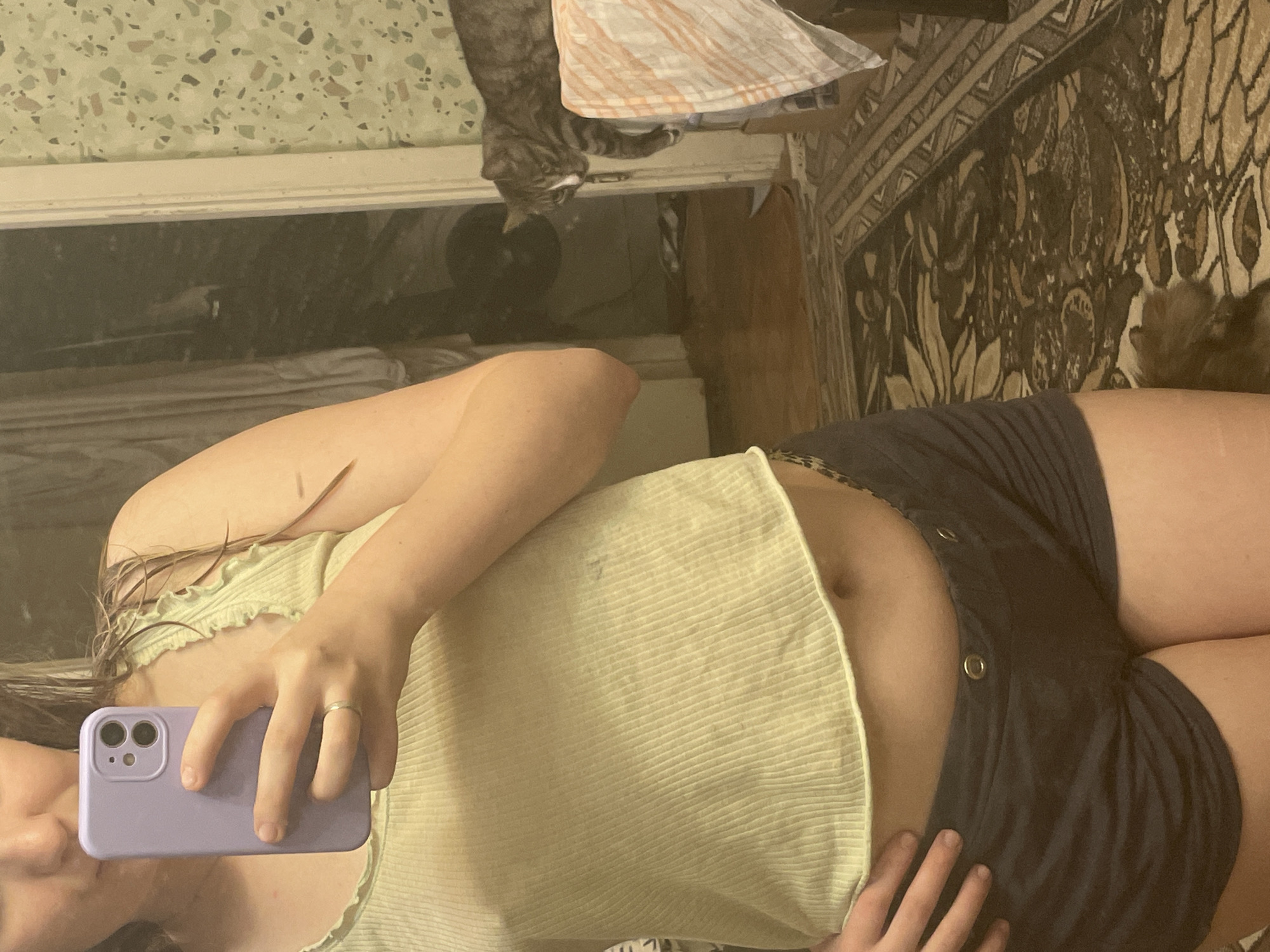 A person stands in front of a mirror, taking a selfie with a purple phone. They are wearing a light green ribbed tank top and dark blue shorts. A cat is visible in the background, lying on a striped blanket. The room appears to be a bedroom, with wood flooring and a patterned rug.