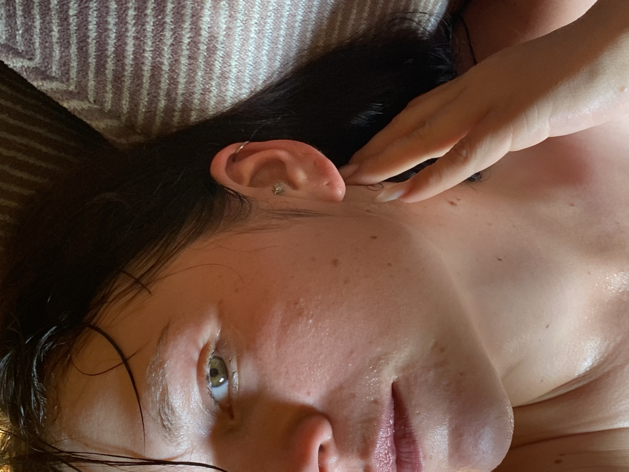 The image is a close-up, eye-level shot of a woman's face and upper neck, with her right hand touching her neck. She has dark hair that is partially wet and falling across her face and shoulders. Her right eye is visible, appearing green or hazel. She is wearing a small stud earring in her right ear, with a hoop earring in her upper ear. Her skin appears slightly shiny, possibly from sweat or product. A portion of a striped fabric, possibly a towel or blanket, is visible in the upper right background. The image is undated and its location in Фоки, Russia, is not visually evident.