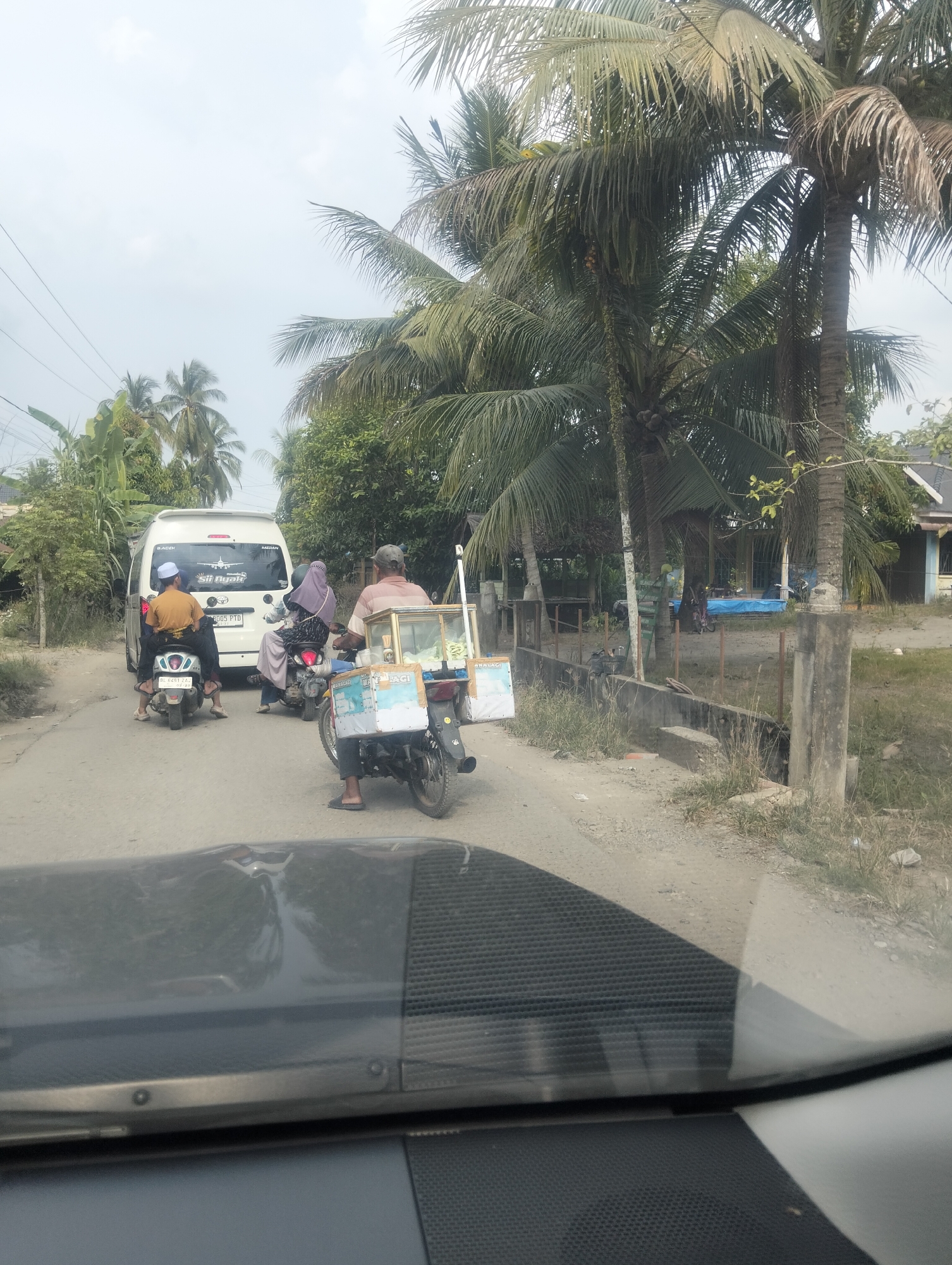 The image depicts a street scene in Teupinreudeup, Indonesia, under an overcast sky, suggesting a warm and humid day. In the foreground, the reflection of palm trees and the sky is visible on the windshield of a vehicle.
The central focus of the image is a traffic jam on a dirt road. A white van is stopped, with text on its rear that appears to be a logo with an airplane and the words "Sili Ayah". Below that, there is a license plate reading "B 0005 FTD".
Several motorcycles are also present in the traffic. One motorcycle in front of the van carries a man wearing an orange shirt and a white cap. Behind the van, two motorcycles are stopped. The first motorcycle has two people on it, a woman in a purple hijab and a man. The second motorcycle, closer to the foreground, is operated by a man wearing a striped shirt and a cap, and it is pulling a cart filled with what appears to be ice cream or similar frozen goods. The cart has text on its side, including the word "JACI" which is partially visible.
The setting is rural, characterized by dense vegetation, including numerous palm trees, and simple buildings with thatched or tiled roofs in the background. A concrete fence runs alongside the road. The overall atmosphere suggests a typical day in a tropical, possibly coastal, Indonesian village. The activity is simply that of vehicles waiting in traffic.