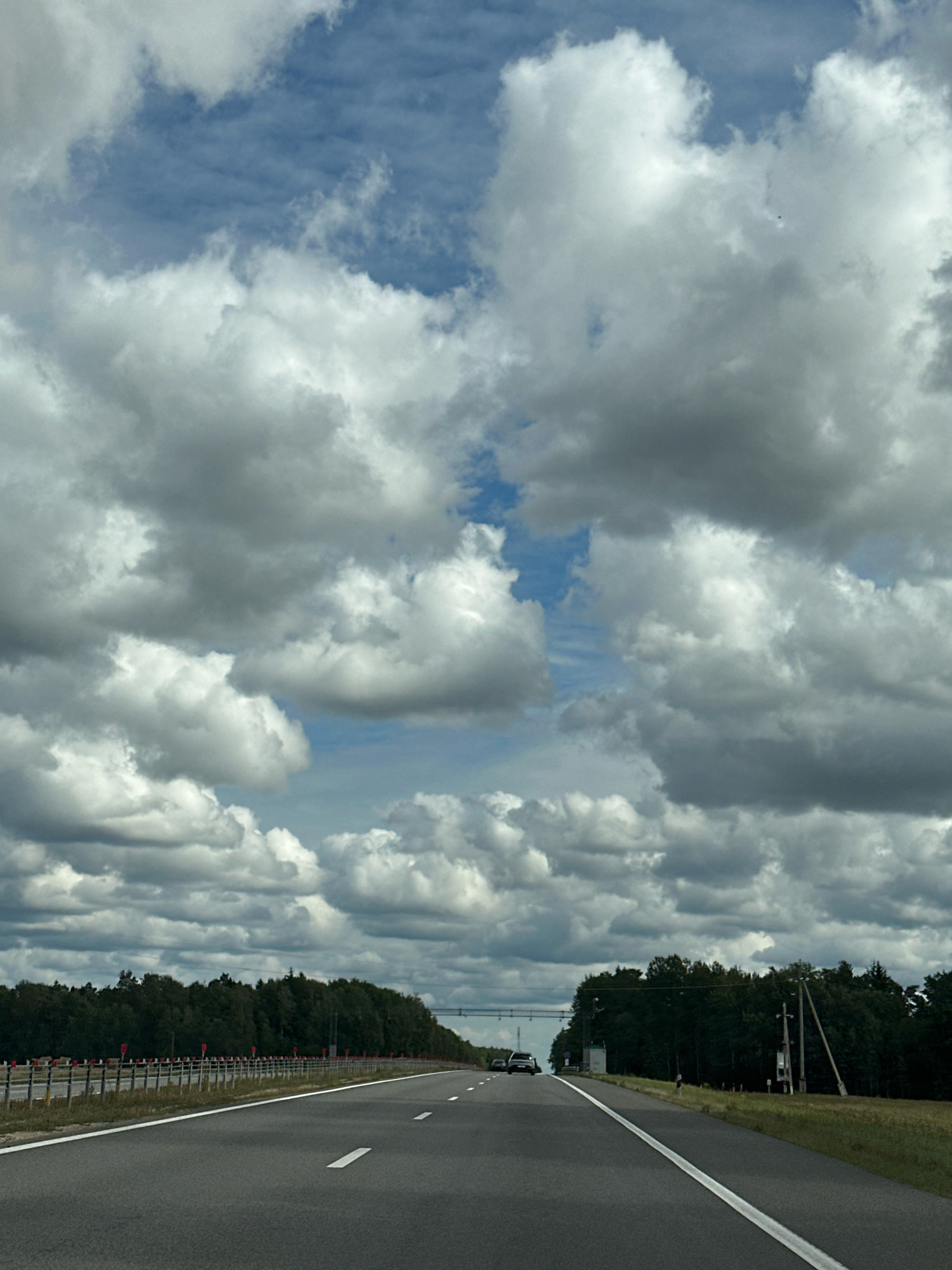 The image shows a highway in Pukhavichy Rural Council, Belarus. The perspective is from a vehicle driving on the road. The sky is filled with dramatic, dark clouds. On the left side of the road, there is a forest with a safety barrier along the edge of the highway. On the right side, there are also trees, and utility poles are visible. Two vehicles are present on the highway ahead, moving in the same direction as the viewer. The road has clear white lane markings. The overall scene depicts a journey on a highway under a cloudy sky, likely during the daytime.