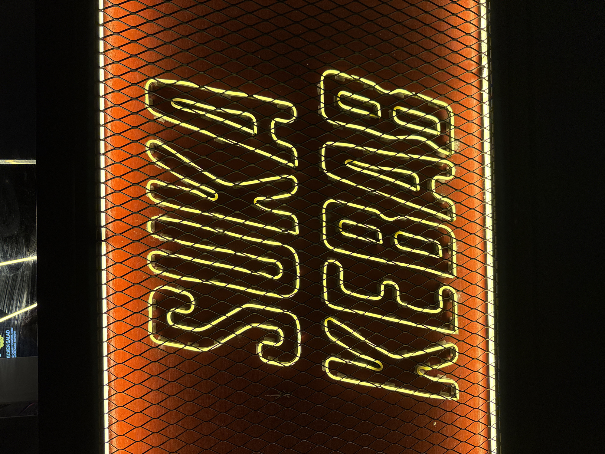 The image shows a neon sign for "Suka Kebab" mounted on a dark-colored wire mesh panel over a reddish-orange background. The sign is written vertically. The photo was taken indoors, possibly in the Ubud District, Indonesia. Part of a menu board is visible on the left.