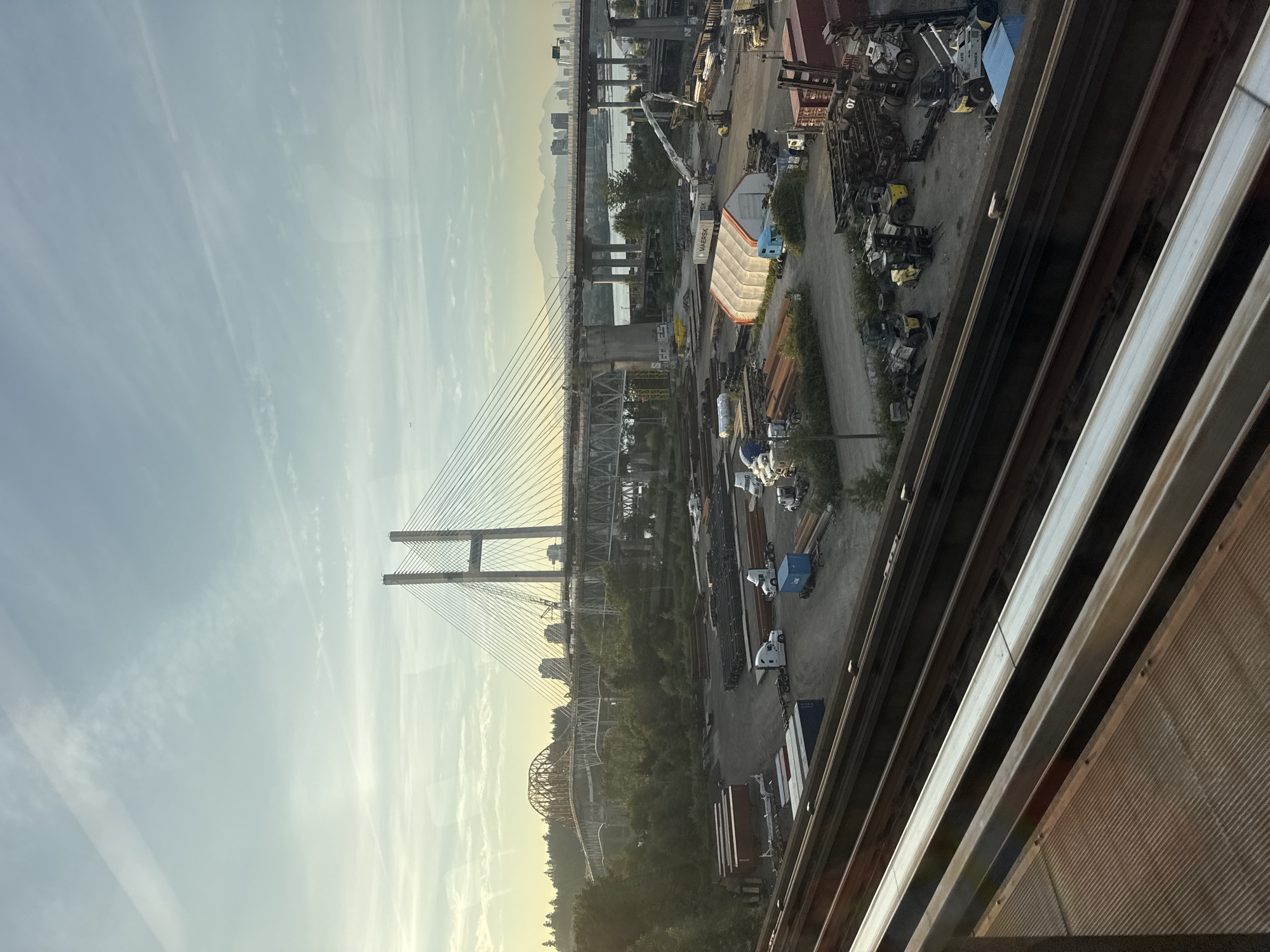 The image depicts a view of the Alex Fraser Bridge in Surrey, Canada, on a bright, clear day. The foreground shows multiple parallel train tracks, with the closest one being a dull metal with a textured surface. Beyond the tracks, a large industrial area is visible, characterized by a variety of shipping containers, construction materials, and heavy machinery.
Several white semi-trucks are parked in the industrial area, some with trailers. Forklifts and other industrial vehicles are also present. A large, white tent with an orange trim is set up in the center of the industrial yard. In the mid-ground, the Alex Fraser Bridge dominates the landscape, a modern cable-stayed bridge with two tall towers. Another older, truss bridge is visible in the distance to the left of the Alex Fraser Bridge.
The background reveals a treeline and a cityscape with several buildings, suggesting an urban or semi-urban environment. Mountains are faintly visible in the far distance. The sky is a pale blue with thin, wispy clouds. The overall impression is of a busy industrial zone adjacent to significant transportation infrastructure, under a bright sky. No people are clearly discernible in the image.