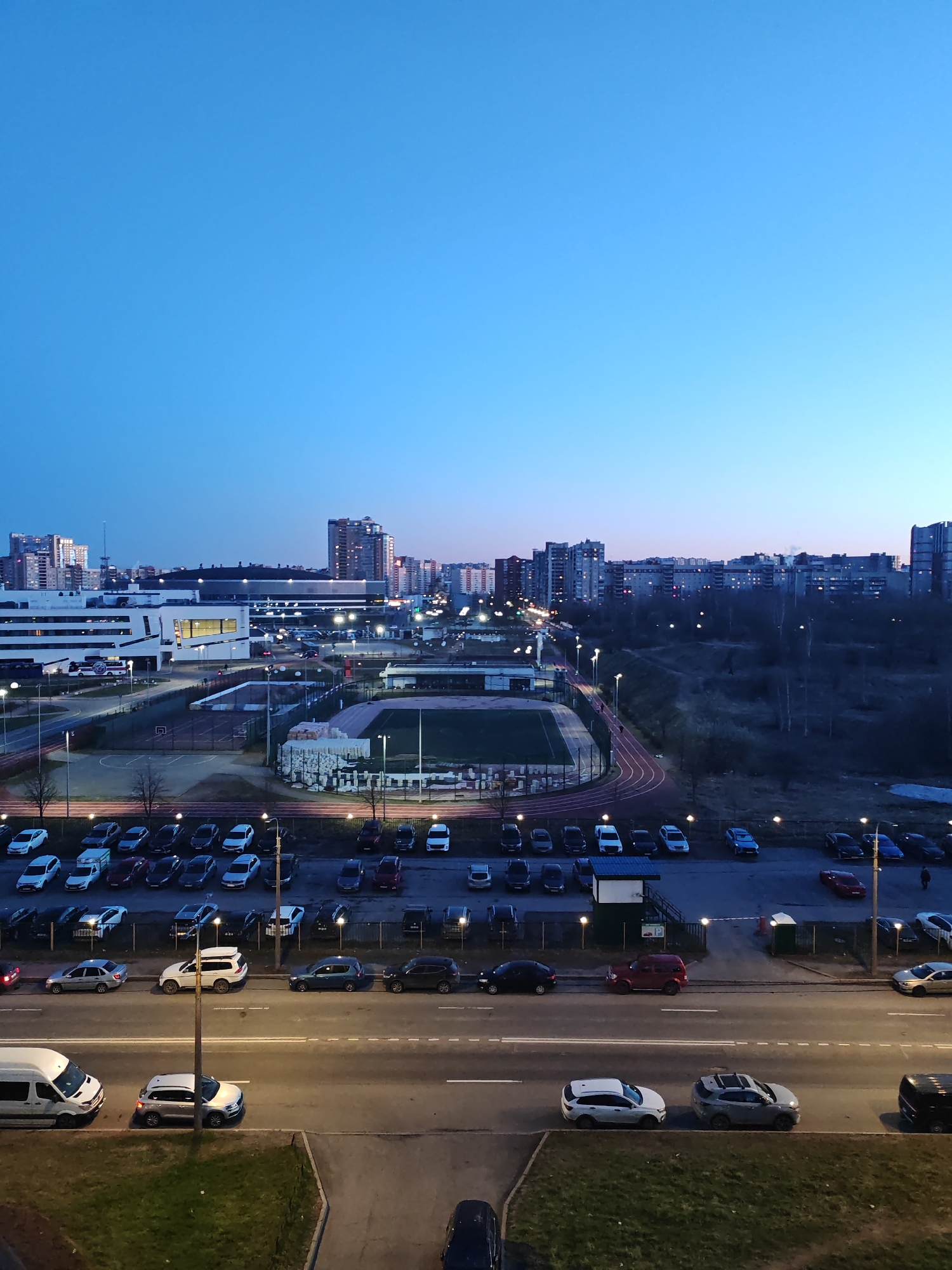 The image shows an elevated view of a cityscape at dusk, with a focus on a sports complex and surrounding urban area. The scene appears to be in Saint Petersburg, Russia, based on architectural styles and general urban layout.
In the foreground, a street with multiple parked cars and a few cars driving along the road are visible. A parking lot adjacent to the street is filled with numerous vehicles.
The central area contains a sports complex featuring a soccer field surrounded by a red running track. Bright lights illuminate the field. There is a complex of interconnected buildings and structures.
The background reveals residential buildings and taller structures, indicating a densely populated urban environment. The sky transitions from light blue to a darker shade, reflecting the approaching night. Trees are present but sparse.
