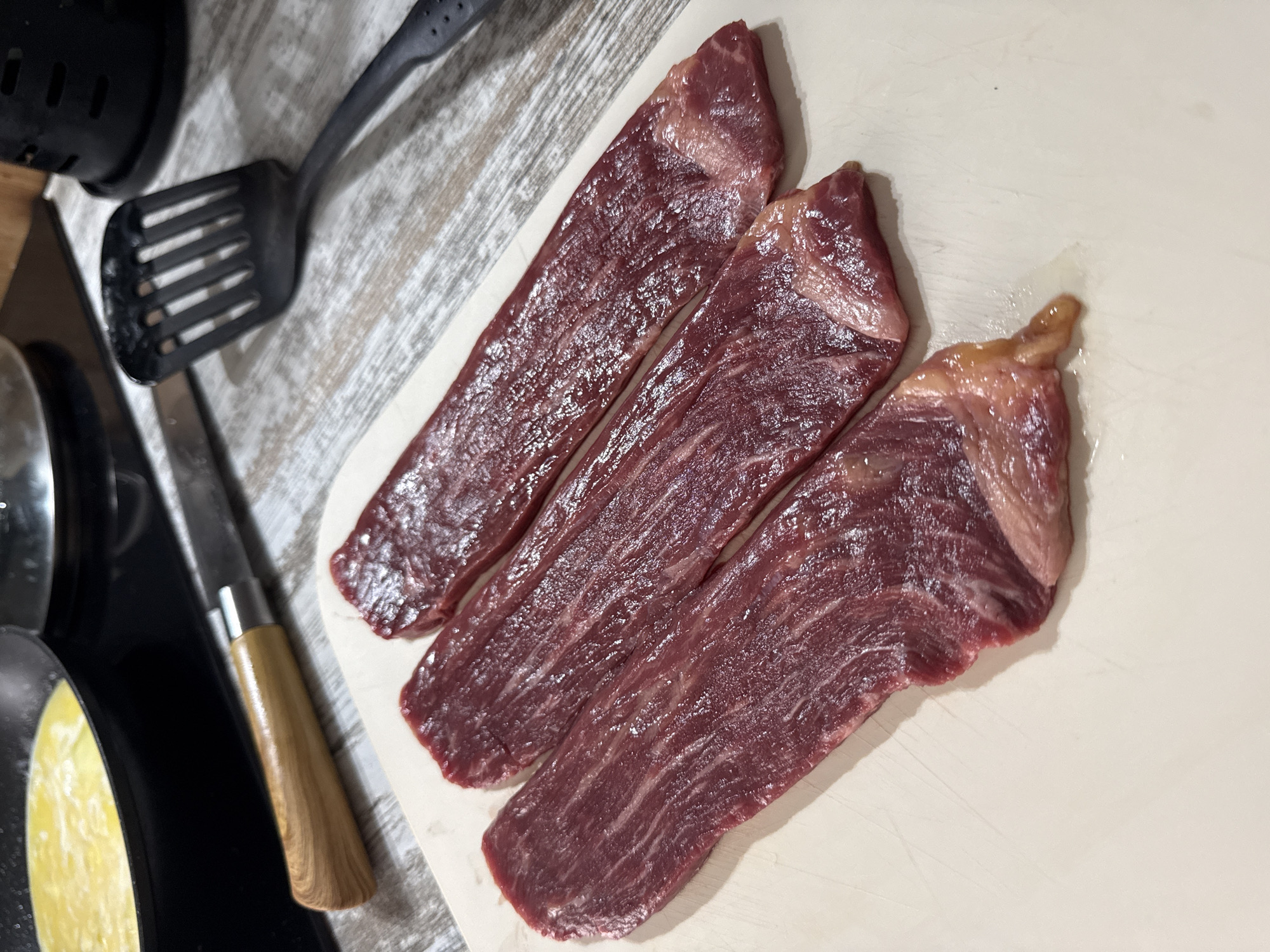 The image shows three raw, elongated cuts of beef lying on a light-colored cutting board. A wooden-handled knife and a black spatula are visible to the left of the cutting board. A black pan is partially visible next to the knife. The image does not contain information about the location of Rostov-on-Don, Russia.
