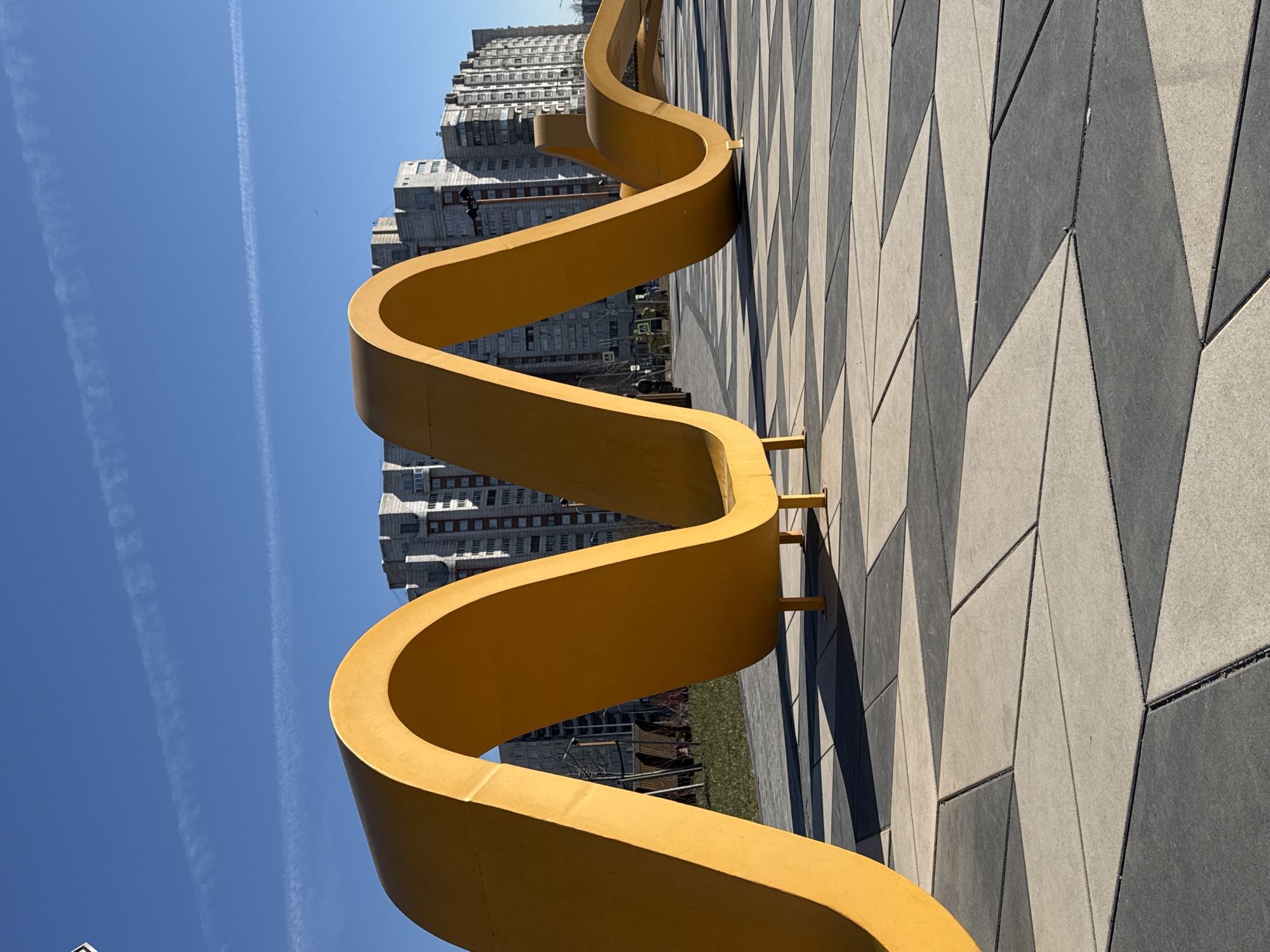A low-angle, medium shot shows a curved, yellow sculpture installed on a patterned walkway against a backdrop of an urban environment under a clear, blue sky.
The sculpture, resembling a stylized sine wave, is rendered in a bright, matte yellow color. It rests on small yellow supports, which attach the structure to the ground. The walkway beneath the sculpture is constructed of large, geometric concrete pavers in alternating shades of light gray and dark gray, arranged in a zig-zag pattern that creates a visual texture.
Beyond the sculpture and walkway, a multi-story building is visible, constructed with a modern architectural style. The structure appears to have a mix of light and dark gray, possibly indicating varying materials or shadows. The facade of the building features numerous windows arranged in a uniform grid pattern.
In the upper left corner of the frame, a blue sky is visible with faint contrails.
The location is the "Park of the 300th Anniversary of Saint Petersburg" in Saint Petersburg, Russia, as per available information.