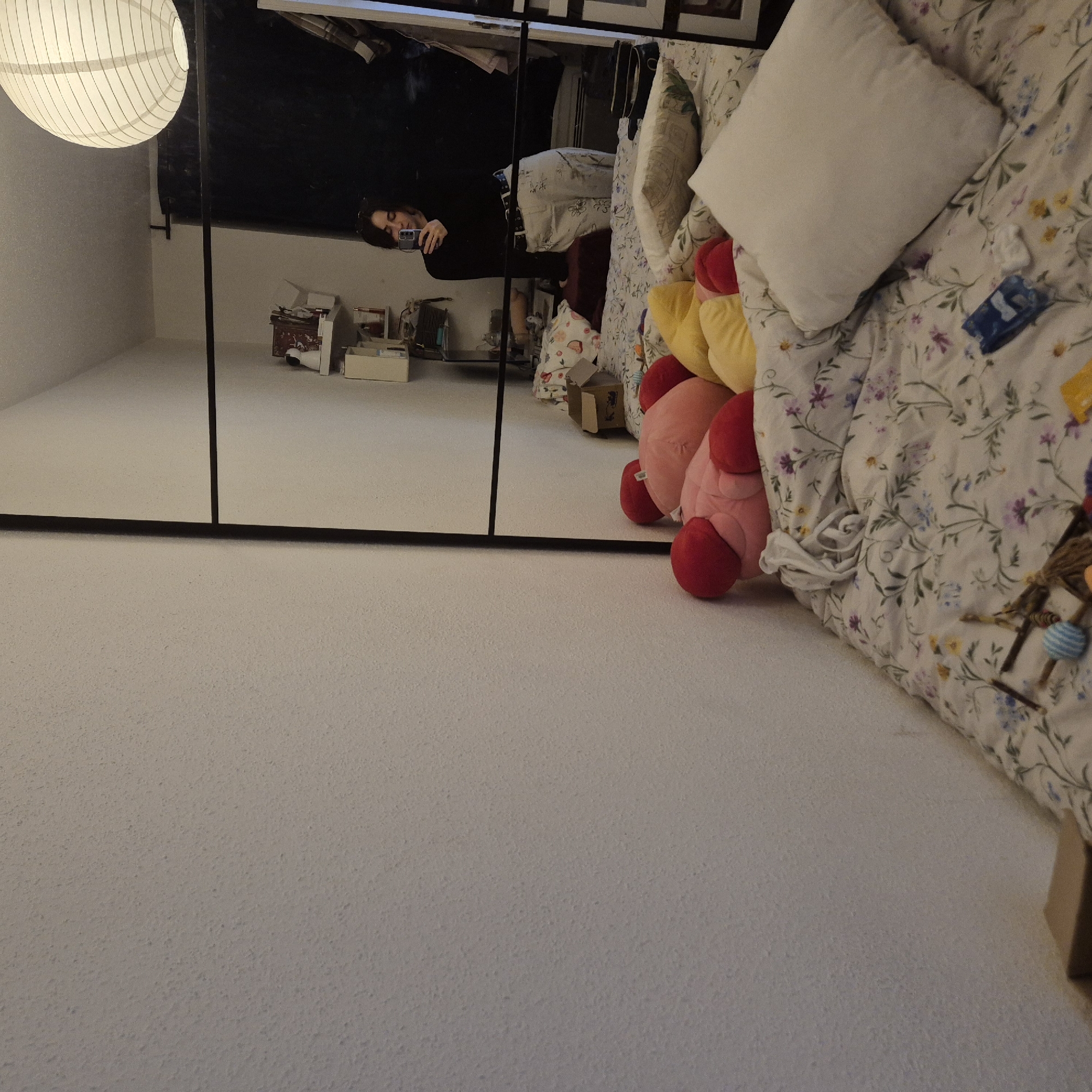 The image shows an interior scene. A black-framed mirror reflects a room with a person holding a phone, presumably taking the picture. The room in the mirror appears to be a bedroom with clothing visible in the background. In the foreground, there is a bed with floral bedding, a white pillow, and several stuffed animals, including pink and yellow ones. The floor is a light color. A paper lantern hangs from the ceiling near the mirror. The location is likely an apartment in Strasbourg, France, given the common architectural styles and typical interior decor found in French residences.