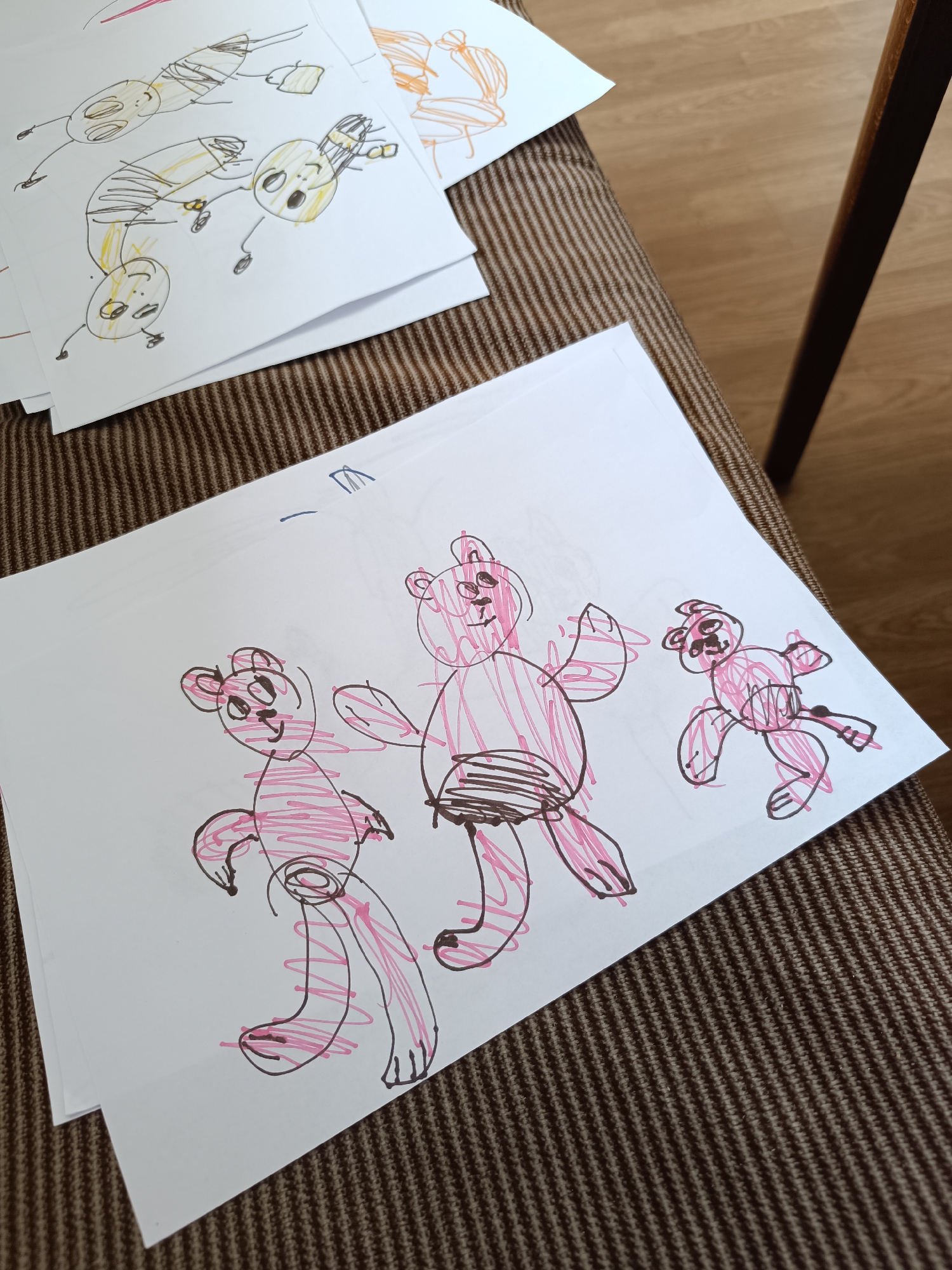 The image shows a close-up of a woven brown surface, possibly a sofa or chair, upon which several pieces of paper are placed. The paper in the foreground contains a child's drawing of three cartoon bears rendered in pink marker with black outlines. The bears appear to be standing upright and possibly gesturing with their arms. Another drawing is visible in the upper left corner, depicting multiple figures drawn in yellow and brown markers. The drawing seems to show abstract representations of people. The scene is set indoors, with a wooden leg of a piece of furniture visible in the background, situated in the city of Unknown, Unknown.