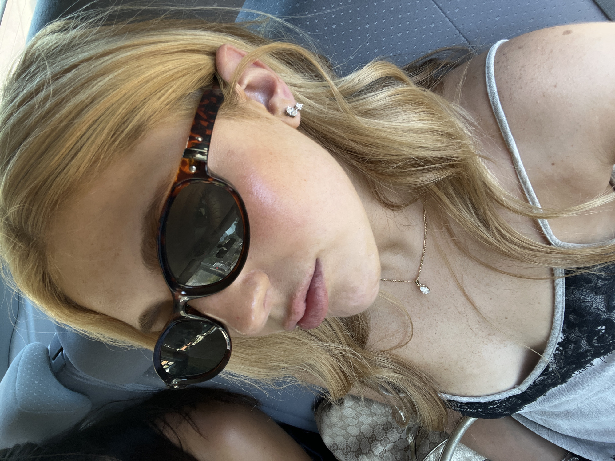 The image shows a woman lying down, possibly in a car, as suggested by the gray seat fabric in the background. She has blonde hair, fair skin, and freckles. She wears sunglasses with a brown tortoiseshell frame. A small silver earring is visible in her ear. She is also wearing a delicate gold necklace with a small pendant. She has on a gray tank top with black lace trim. A patterned purse rests near her. The reflection in her sunglasses reveals a street scene which is consistent with what is known about Antibes, France.