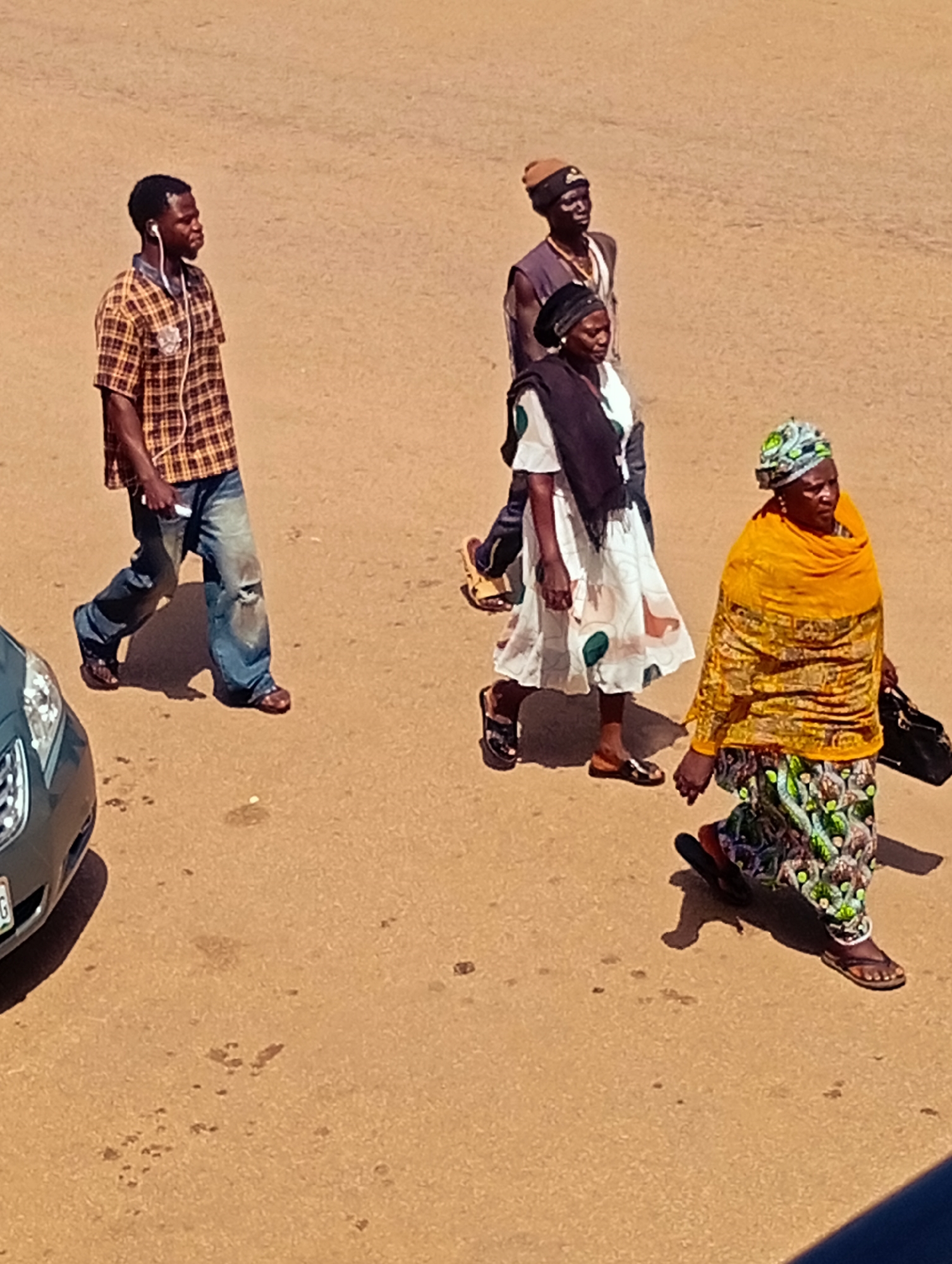 In Bukuru, Nigeria, four individuals are captured walking on a dusty, outdoor surface, possibly a street or plaza. The scene is illuminated by sunlight, casting distinct shadows. On the left, a man in a plaid, collared shirt and distressed denim jeans is walking, his right earbud in. He is moving away from the viewer, towards the right. Behind him, slightly to the right, two individuals are walking together. One, to the left, is wearing a dark headwrap and a white dress with a black shawl draped over their shoulders. To the right of this person and slightly behind, a man wears a dark vest over a light-colored garment and a brown beanie. Further to the right and ahead of the other three, a woman walks briskly, holding a dark bag in her right hand. She wears a vibrant yellow wrap and a patterned skirt, with a green and blue patterned headscarf. Her footwear appears to be sandals. A dark-colored car is partially visible on the far left of the image, with its front end facing the left edge of the frame. The ground is a uniform sandy-brown color, with scattered dark spots and some lighter splotches, suggesting it is a natural or unpaved surface. The overall impression is of a casual, everyday scene unfolding in a Nigerian urban environment.