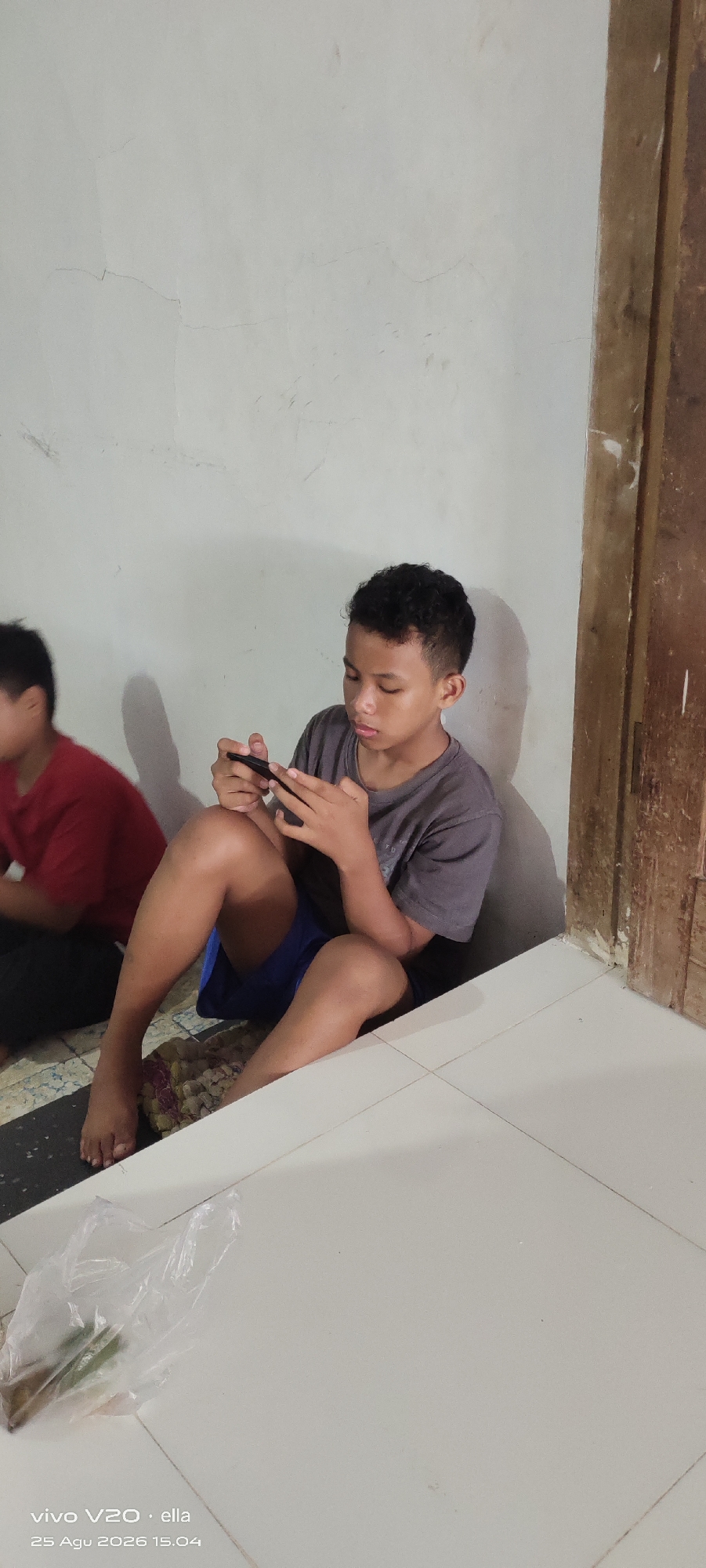 In Simo, Indonesia, a young boy with dark, curly hair is seated on the floor, leaning against a plain, light-colored wall. He is wearing a grey t-shirt and blue shorts, and is engrossed in playing a game on his black smartphone, which he holds with both hands. His legs are crossed, with his right foot visible. To his left, partially visible and out of focus, another young boy in a red t-shirt is also seated. The environment appears to be indoors, with a wooden door frame visible on the right side of the image. The floor is tiled with light-colored tiles, and a plastic bag containing some green items is placed on the floor in the foreground. The wall has some visible cracks. The bottom left corner of the image displays a timestamp: "vivo V20 · ella 25 Agu 2026 15.04".